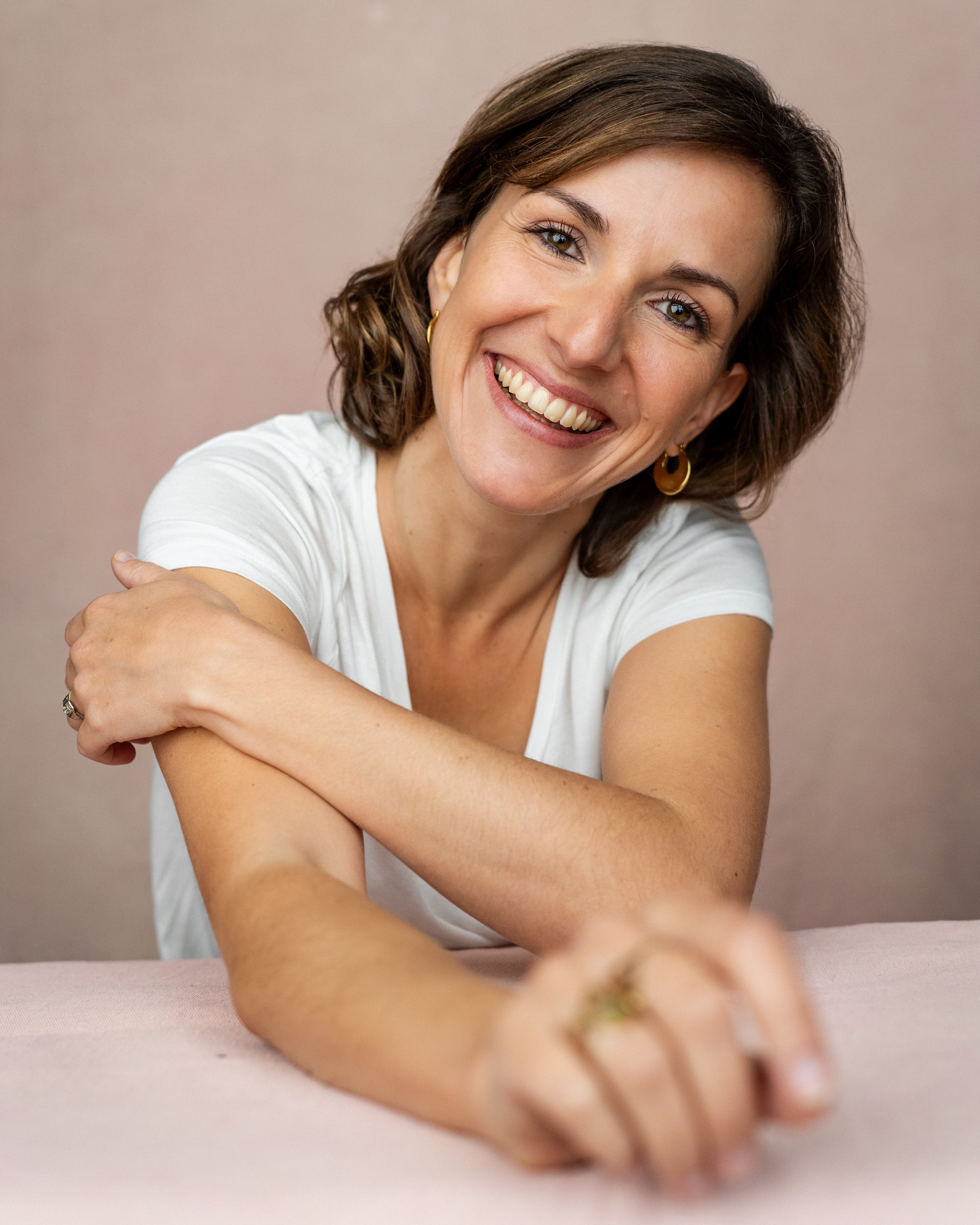 A woman with short brown hair, smiling, wearing a white shirt and gold hoop earrings, leaning forward with her arms crossed on a light pink surface against a plain beige background.