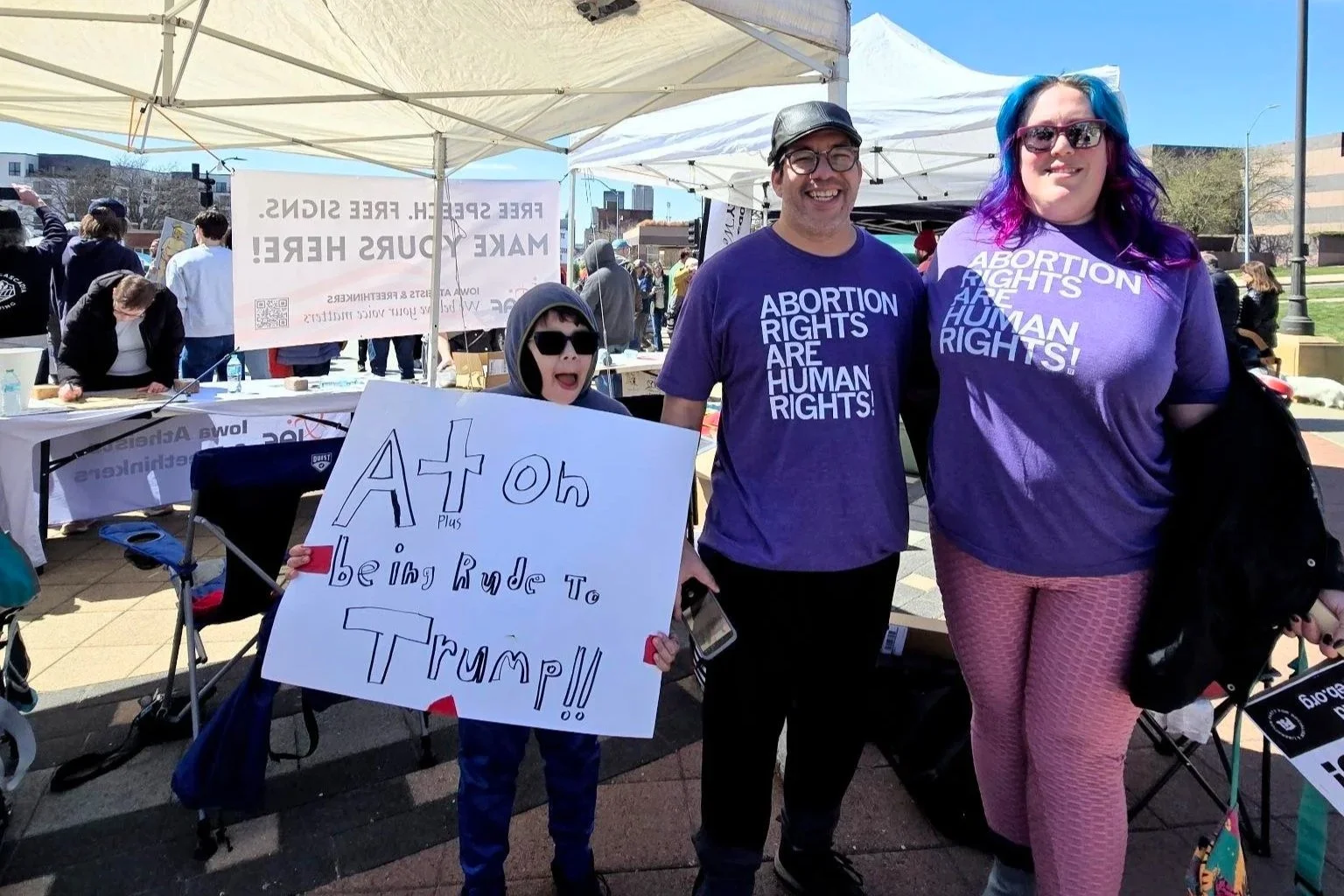 Three people at a No Kings rally, wearing purple shirts that say 'Abortion Rights Are Human Rights,' standing under a white tent. The child on the left holds a homemade sign that reads 'A+ to being rude to Trump!!' in large, colorful letters.