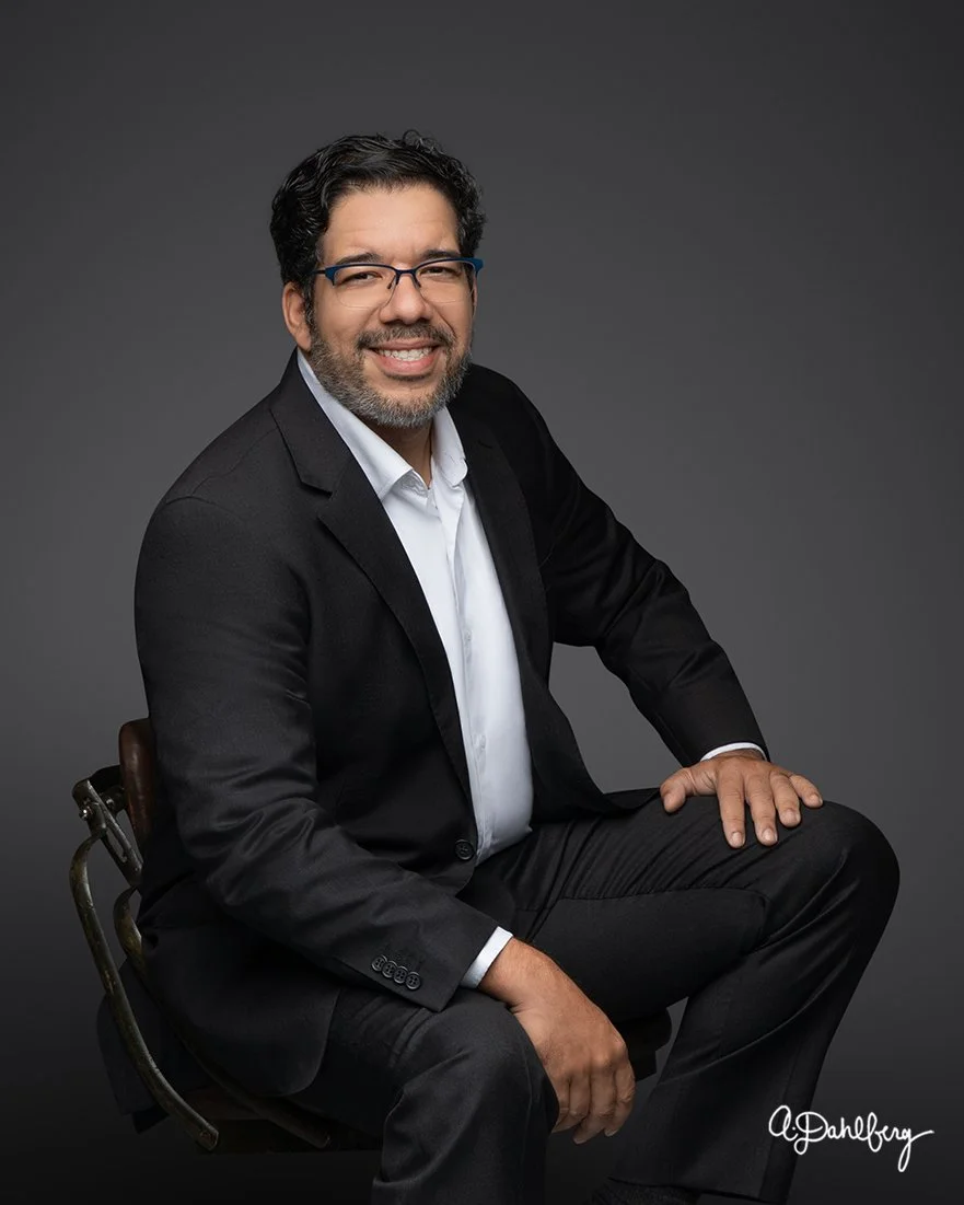 Portrait of Jason Benell. He has dark, curly hair and glasses, wearing a black suit jacket and white shirt, sitting on a chair against a dark gray background, smiling at the camera.