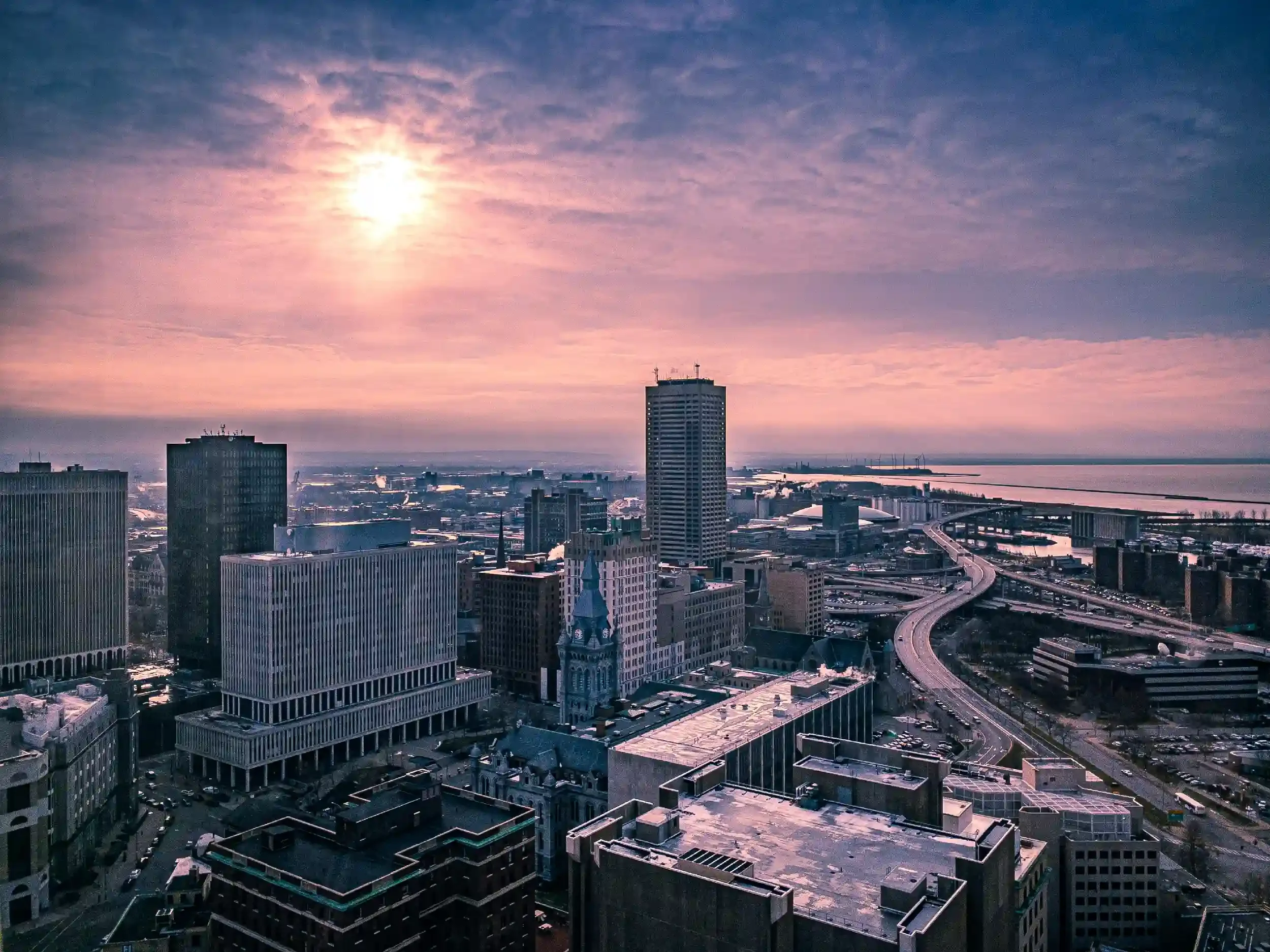 A city skyline at sunset with tall buildings, a river, and bridges under a colorful sky with clouds.