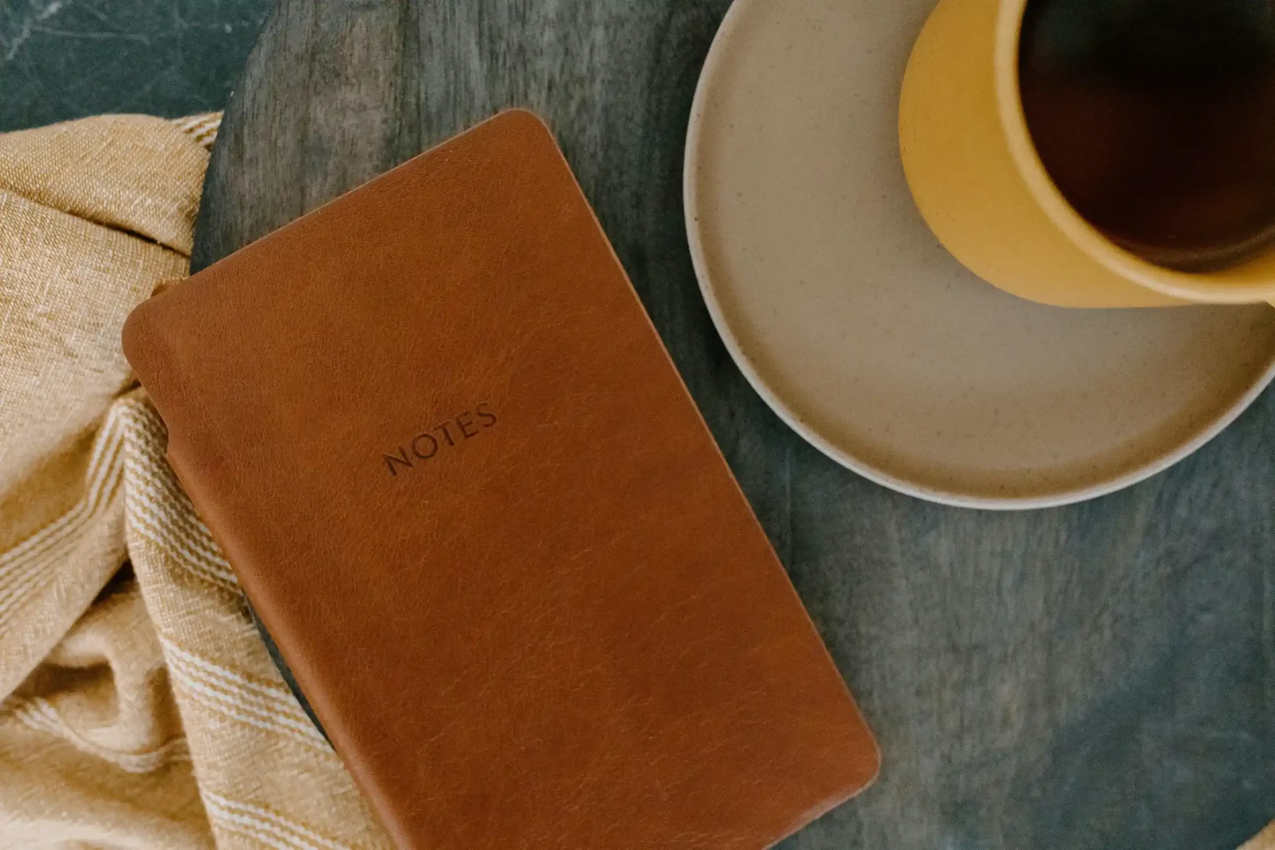 A brown notebook labeled 'NOTES' next to a beige and white striped cloth, a yellow cup with black coffee on a white saucer, and a beige cloth on a dark surface.