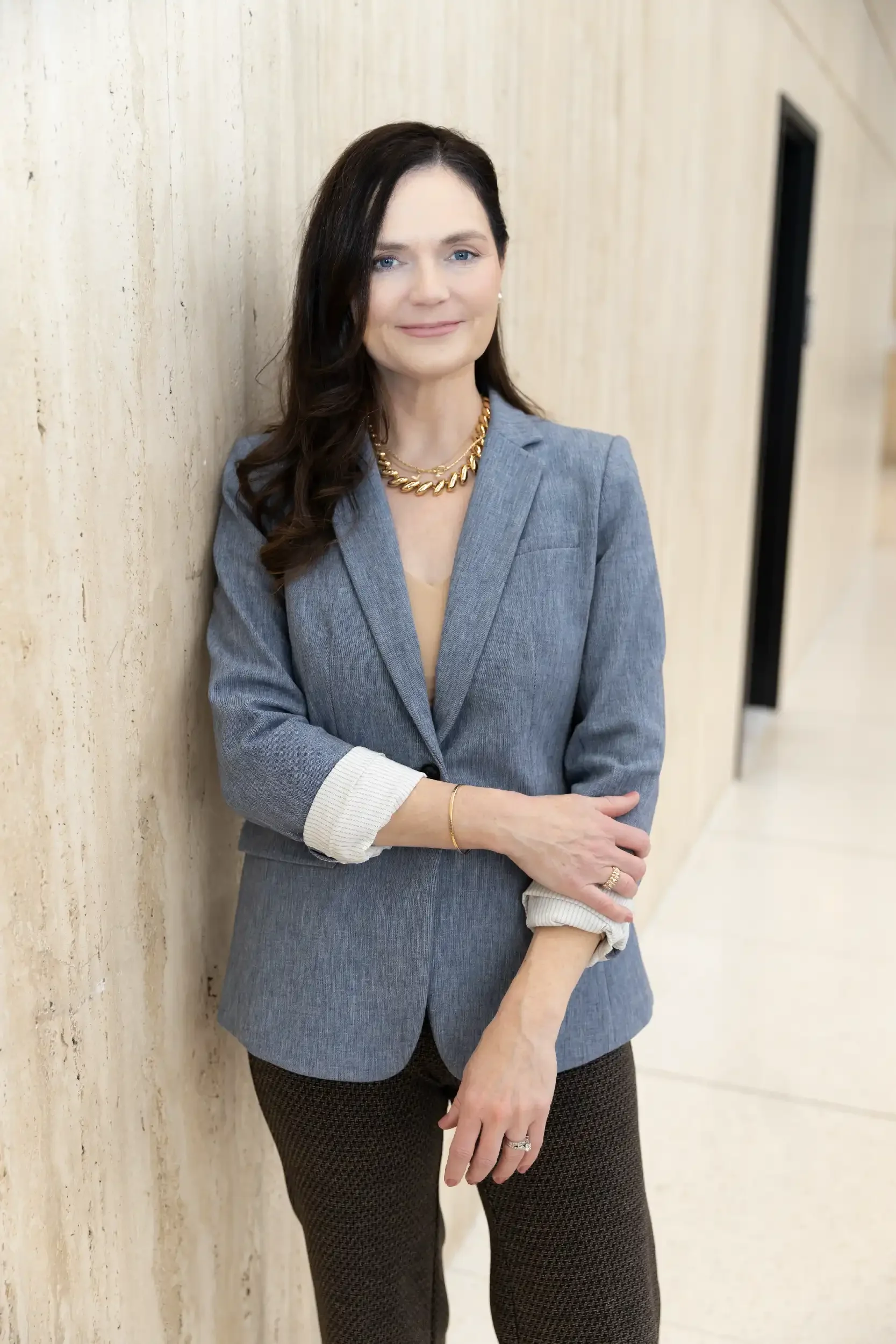 A woman with dark brown hair and blue eyes wearing a gray blazer with white cuffs, a gold necklace, and black patterned pants, standing against a beige textured wall.