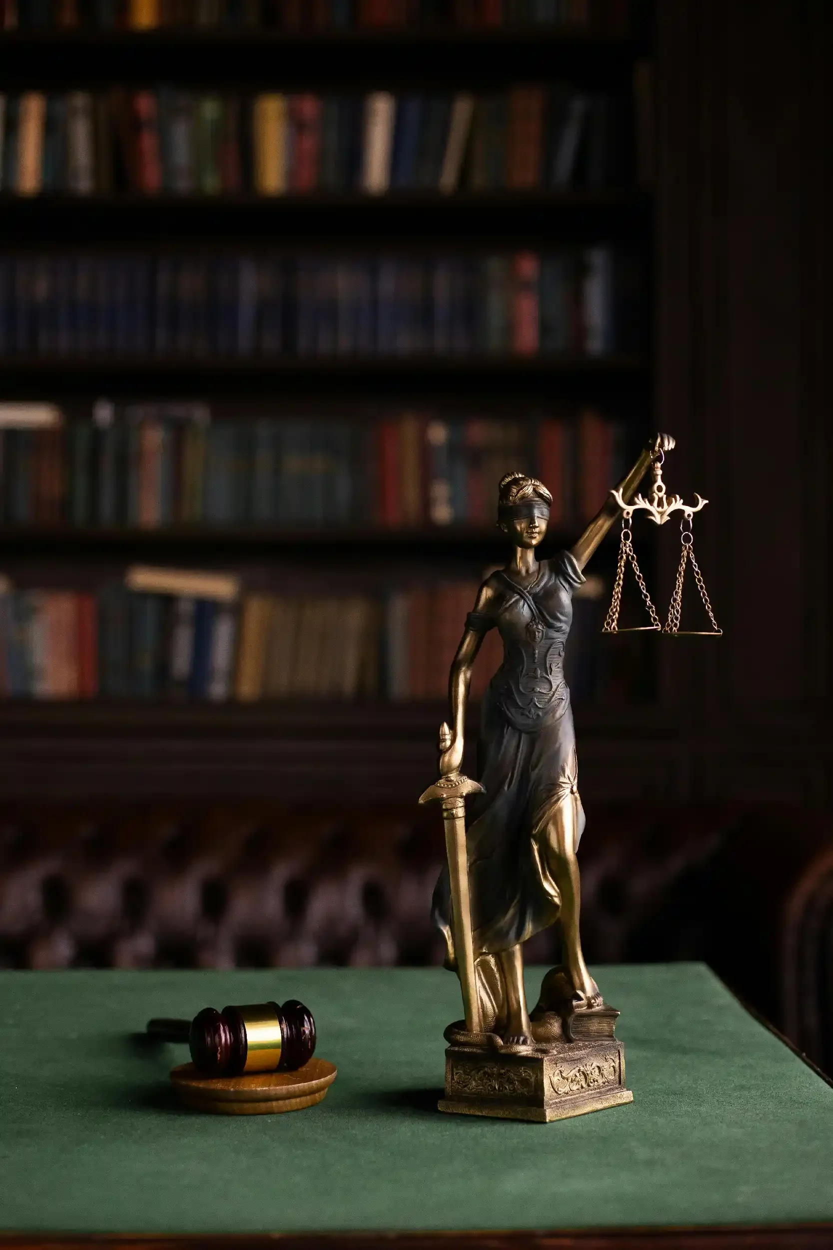 A bronze statue of Lady Justice holding a scale in her left hand, standing on a decorative base, on a green table with a gavel and a ring box, against a blurred background of bookshelves.