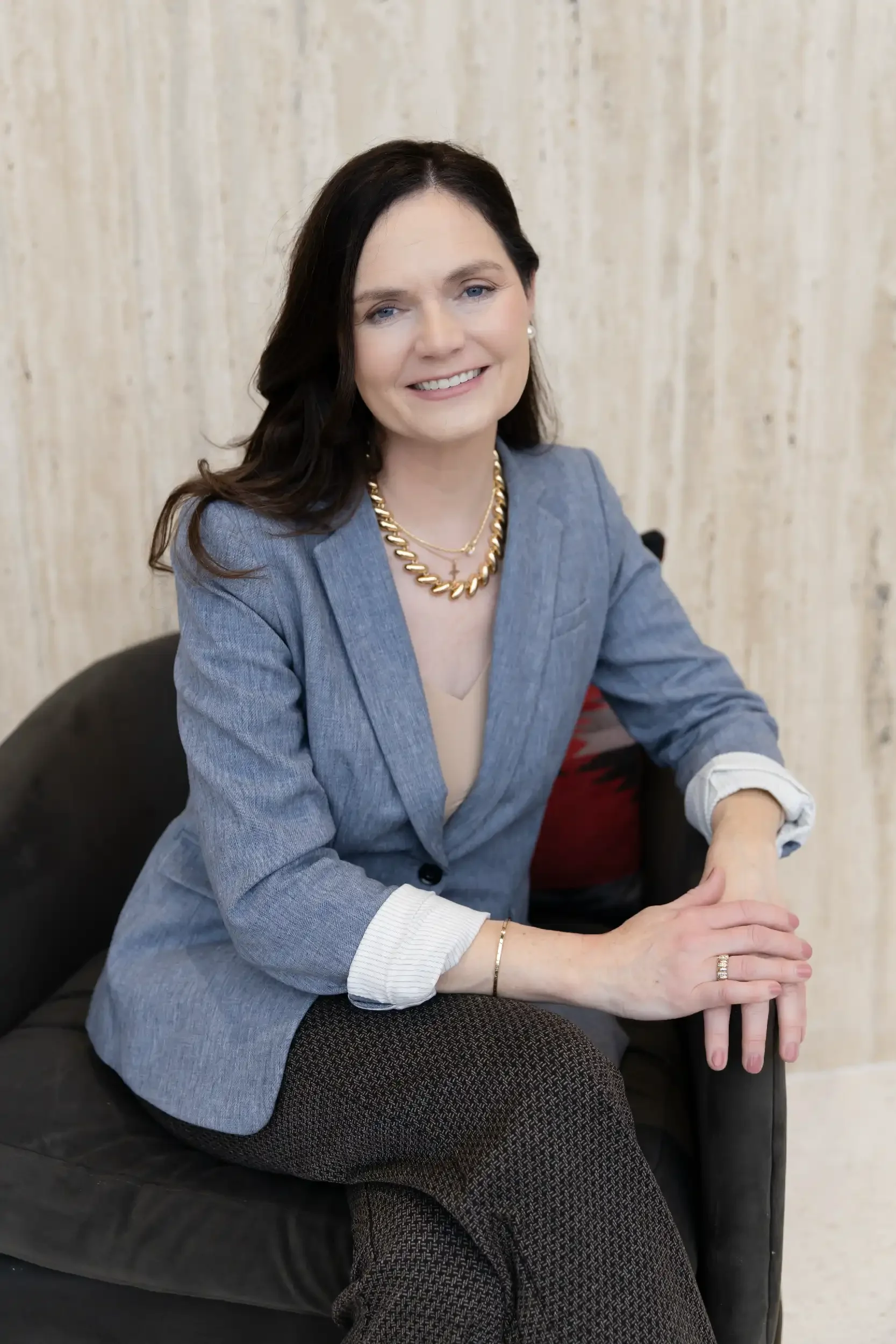 A woman with dark brown hair, wearing a blue blazer, gold chain necklace, and jewelry, sitting on a black couch with her hands clasped, smiling against a neutral background.