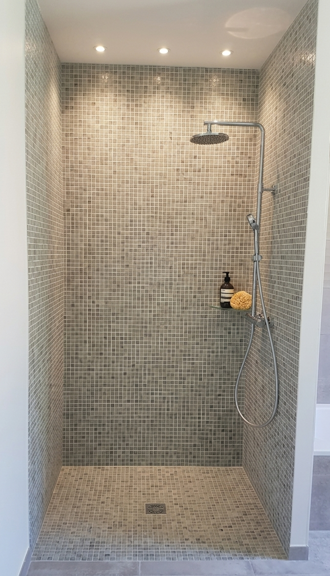 Douche à l'italienne avec carrelage gris, pomme de douche ronde, bouteille de savon et éponge sur une étagère.