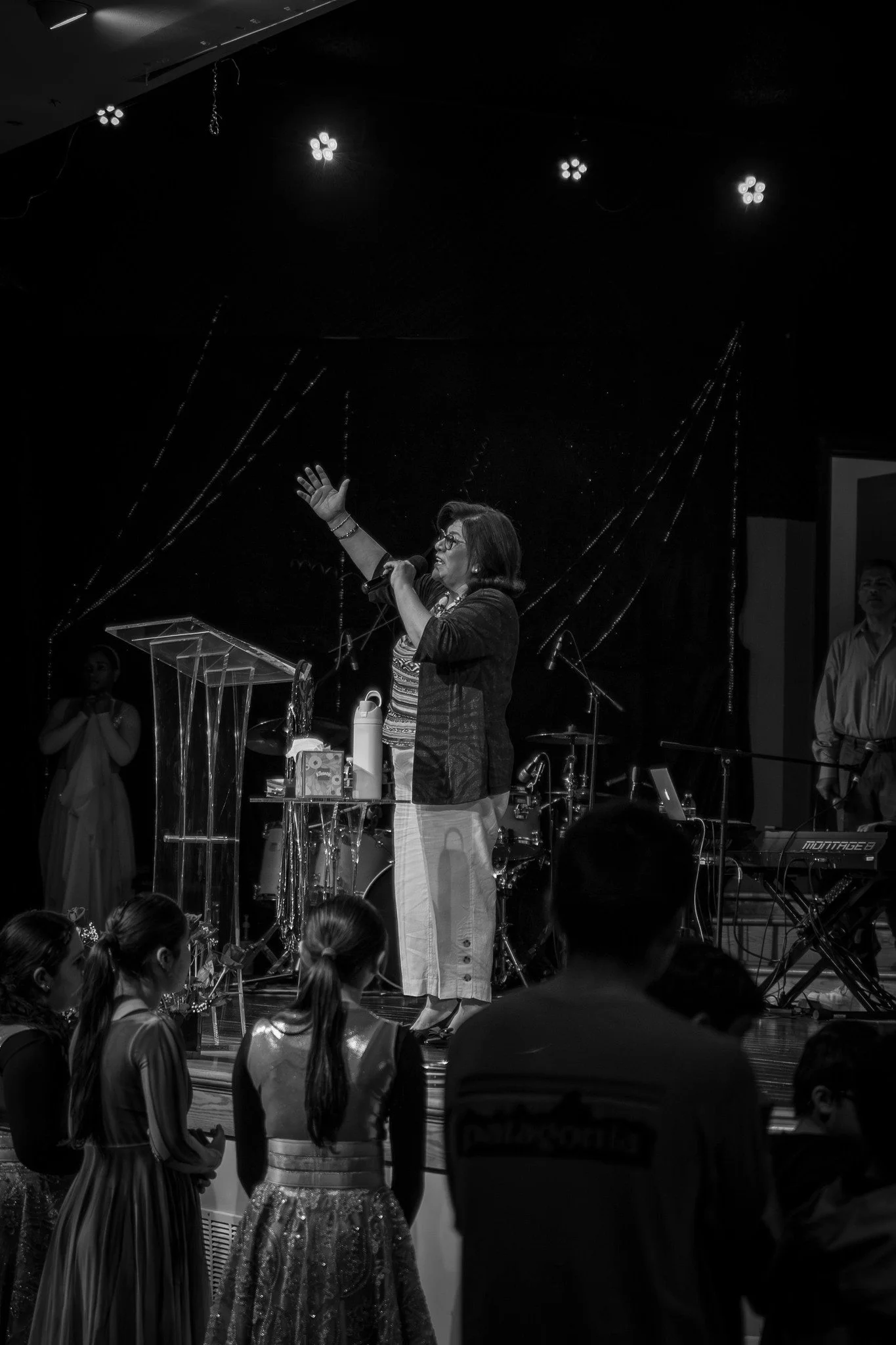 A woman with glasses speaking into a microphone on stage, gesturing with her left hand, surrounded by a music band and an audience.