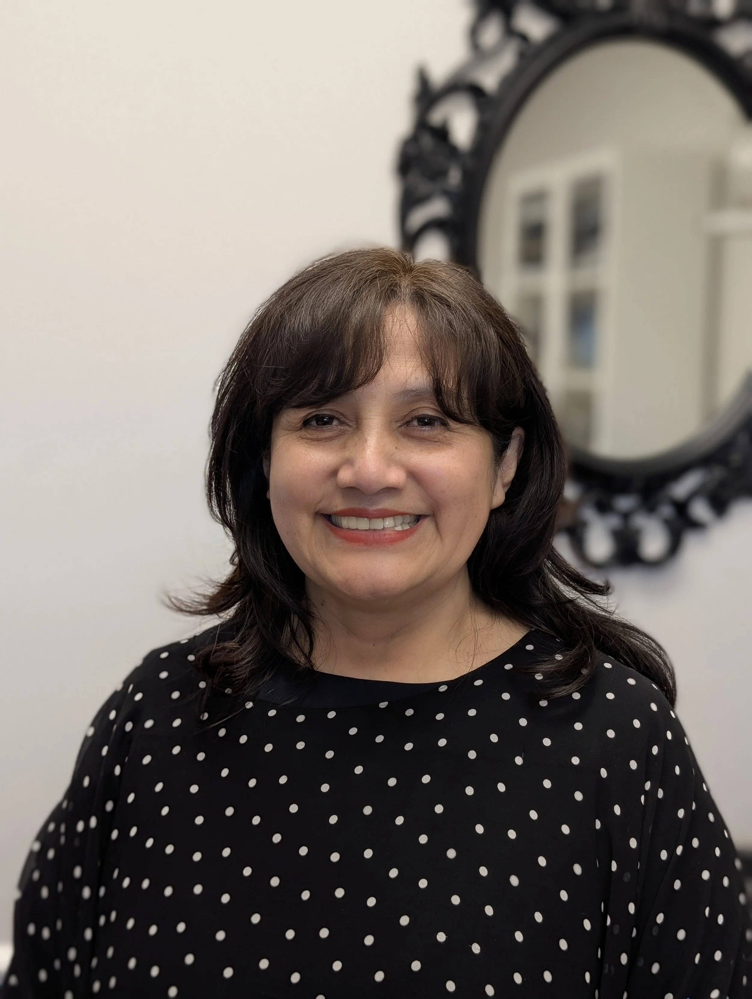 A woman with shoulder-length dark hair and a black dress with white polka dots, smiling, standing in front of a white wall with a black ornate mirror.