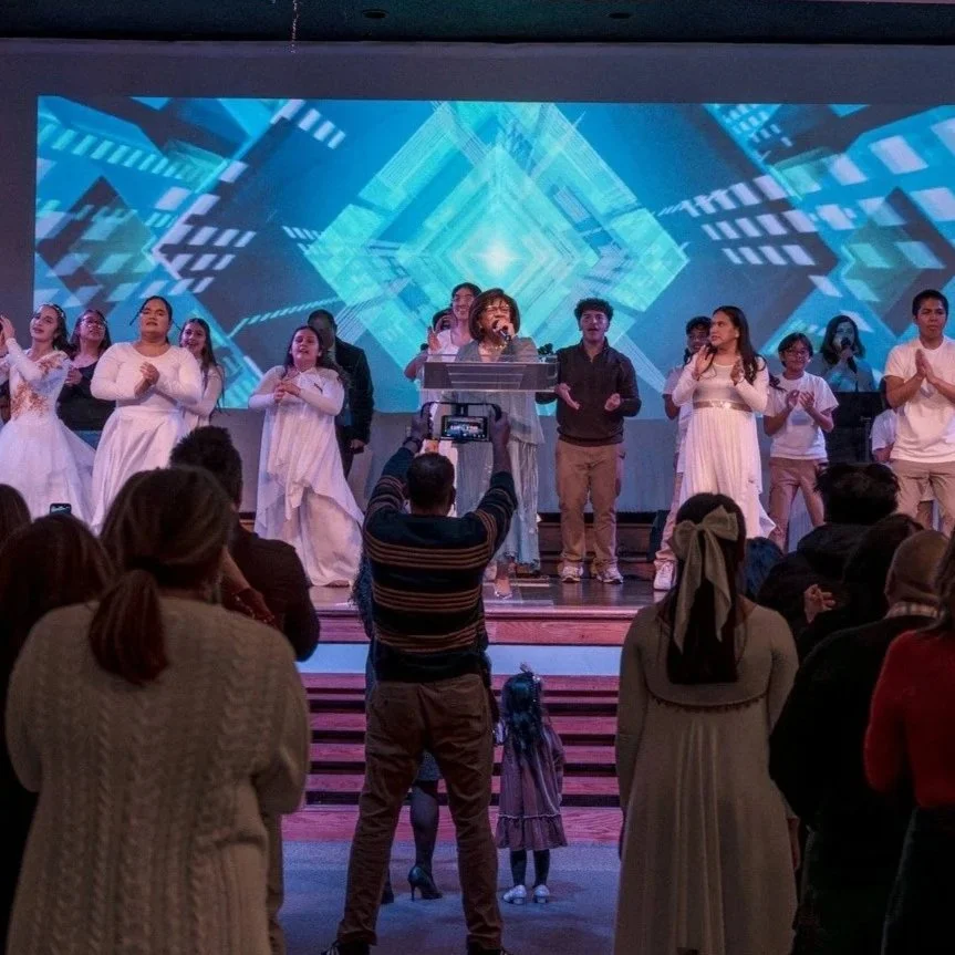 A group of people on stage, some are singing or speaking into microphones, with a large LED screen displaying geometric patterns behind them. The audience is watching and taking photos.