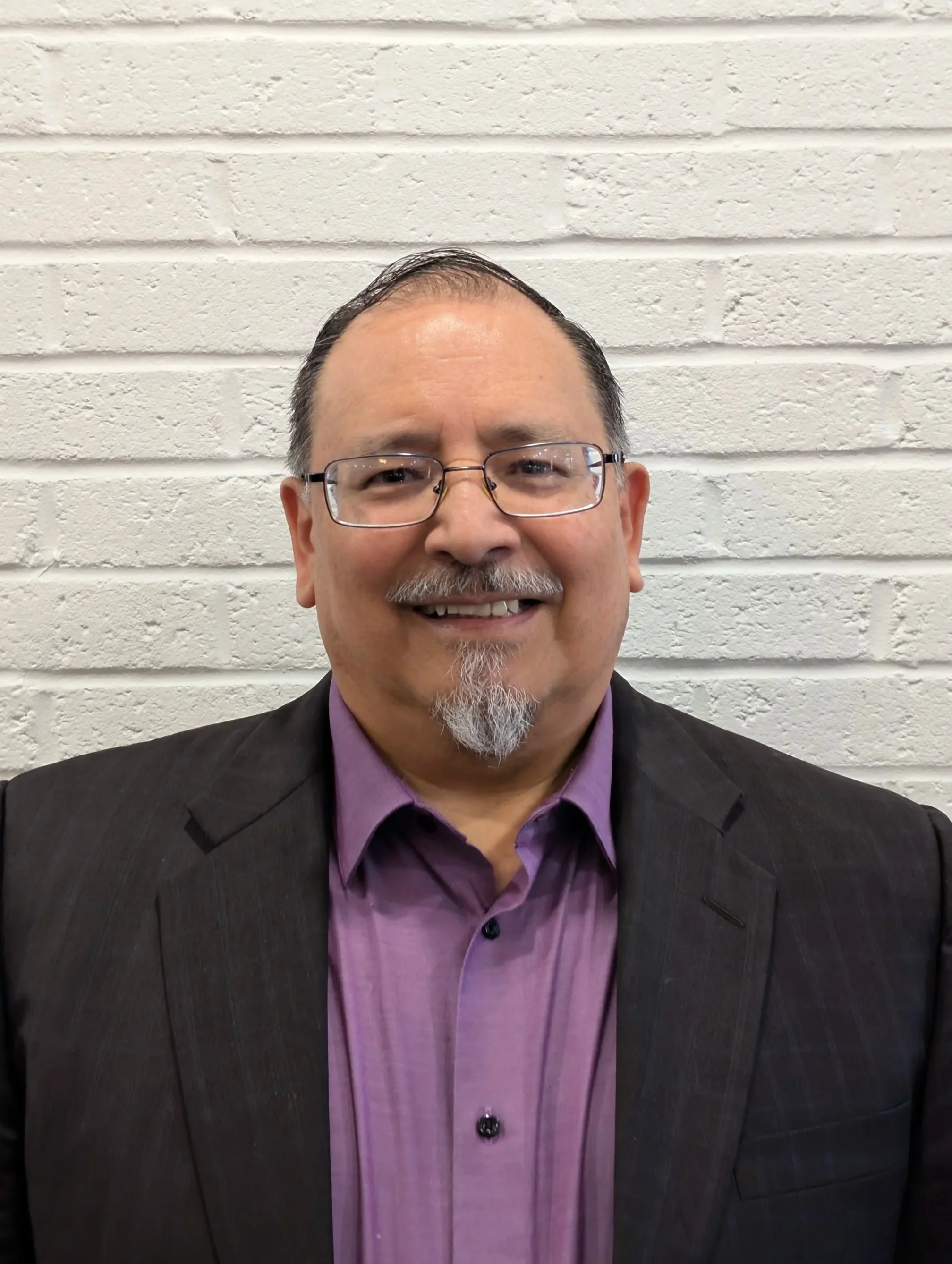 A smiling man with glasses, a goatee, and short dark hair, wearing a purple shirt and dark suit jacket, standing against a white brick wall.