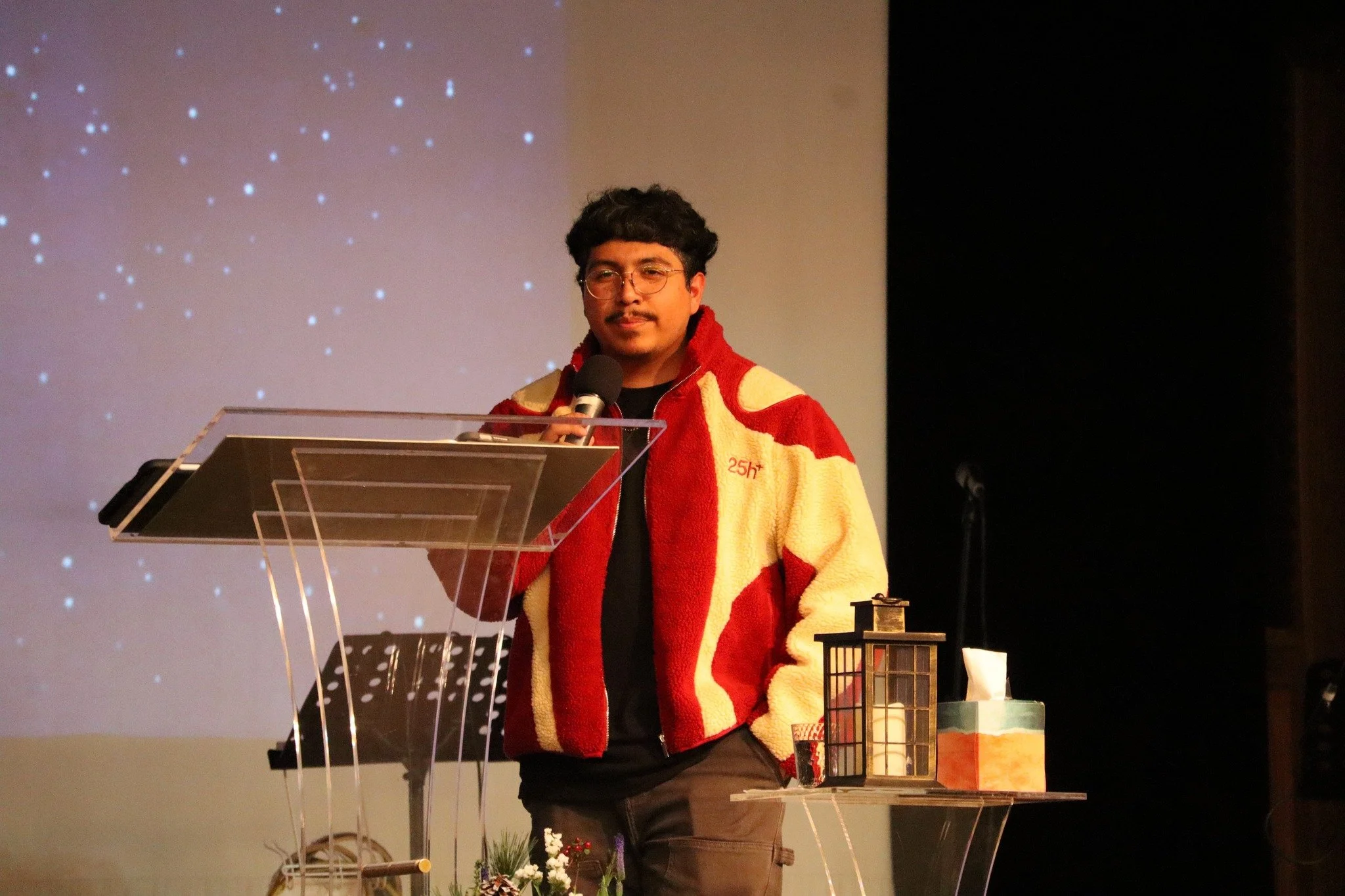 Young man with curly hair and glasses wearing a red and yellow jacket, standing at a podium with a microphone in front of a starry background, speaking or presenting.