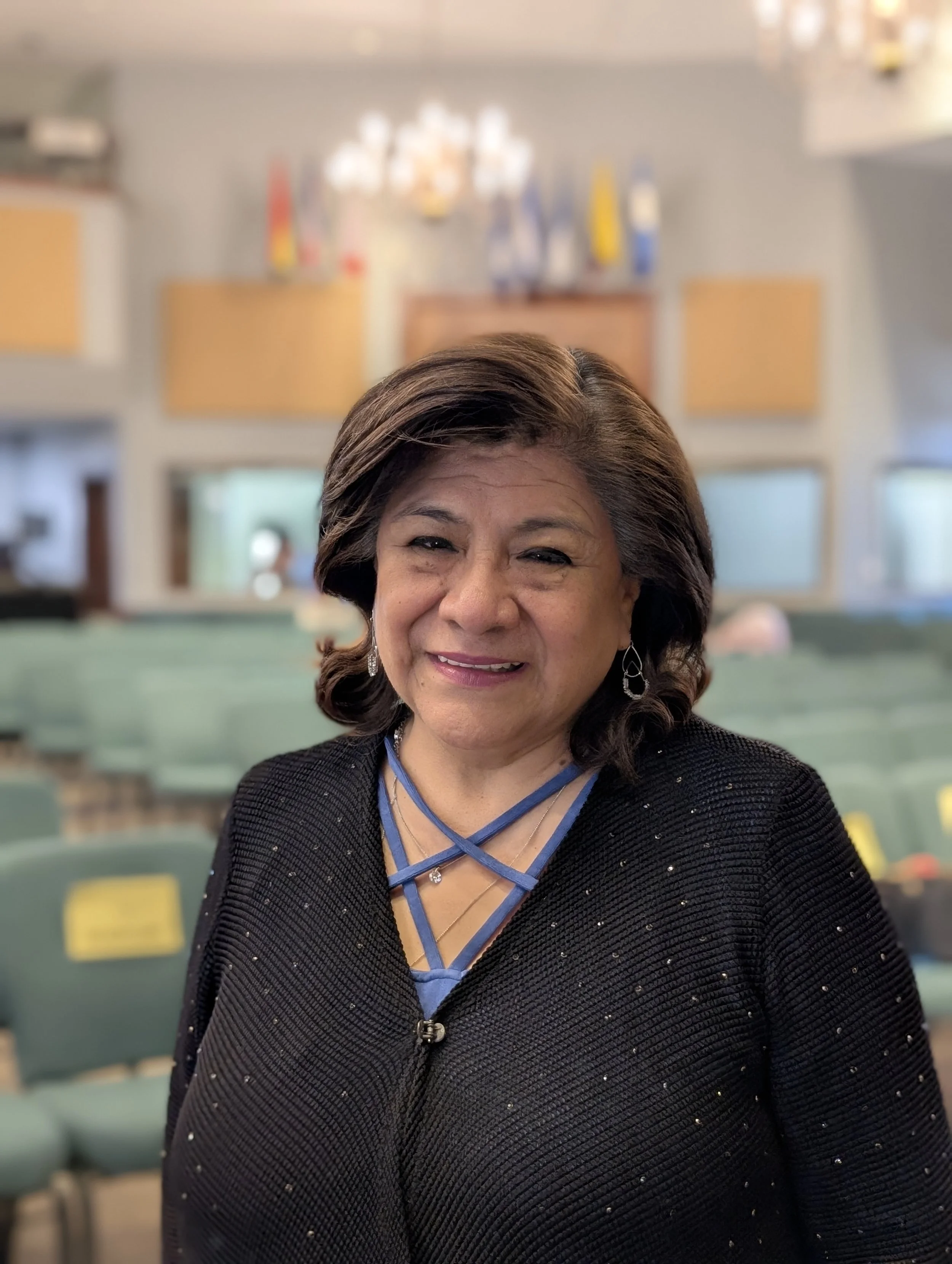 A woman with shoulder-length dark hair smiling in a large room with teal seats and colorful decorations on the wall behind her.