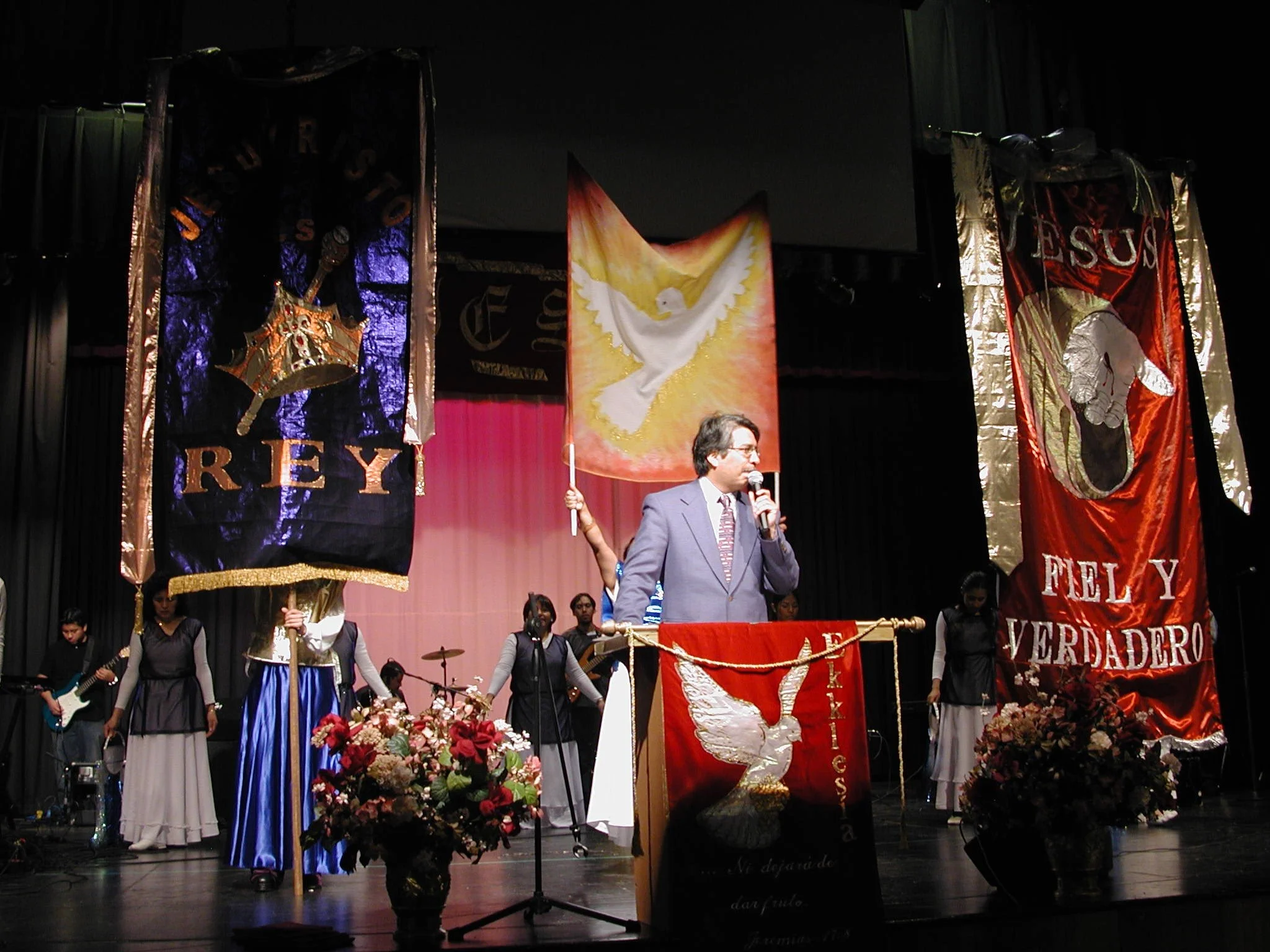 A man in a suit speaking into a microphone on stage, holding a flag with a dove symbol. Behind him are large banners and people holding flags, with flowers at the front of the stage.