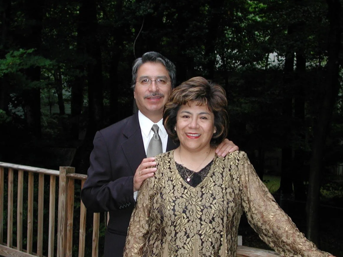 A man and woman posing outdoors on a wooden deck with trees in the background. The man has dark hair, glasses, and is wearing a dark suit with a white shirt and a silver tie. The woman has short, curly hair and is wearing a gold lace dress with black decorative elements and a necklace. They are smiling and standing close together, with the man's hand resting on the woman's shoulder.
