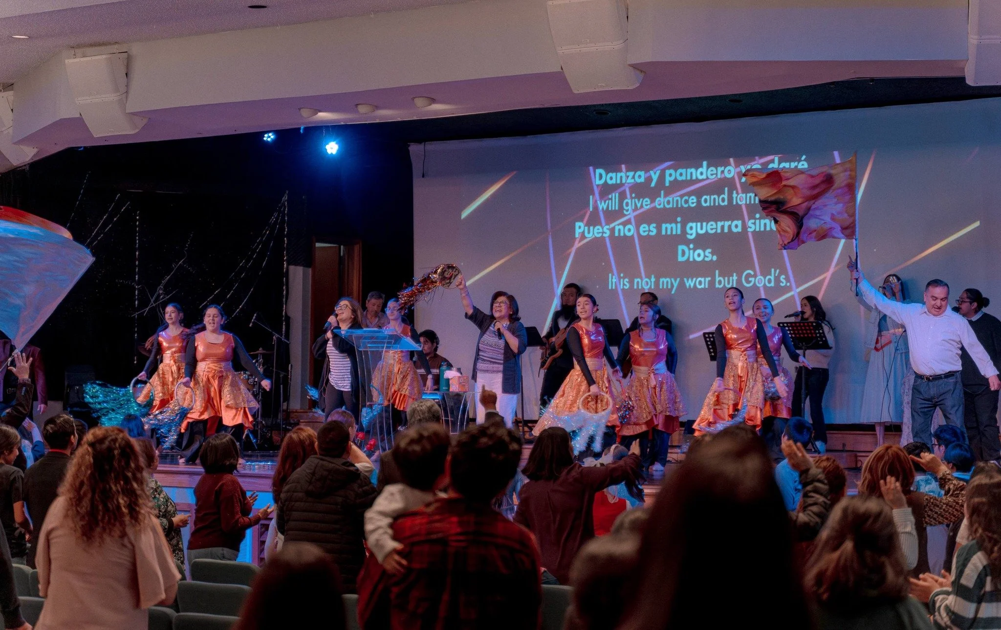 People on stage in a church or auditorium engaged in a performance with dancers in orange costumes, a woman with a microphone, and a man waving a flag, with an audience clapping and singing along, and lyrics projected on a screen behind.