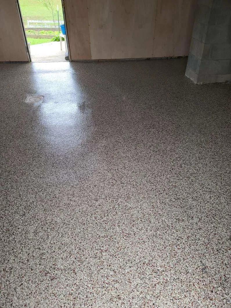 Interior of a room with a speckled epoxy floor, unfinished walls, and an open door leading outside to a grassy area.