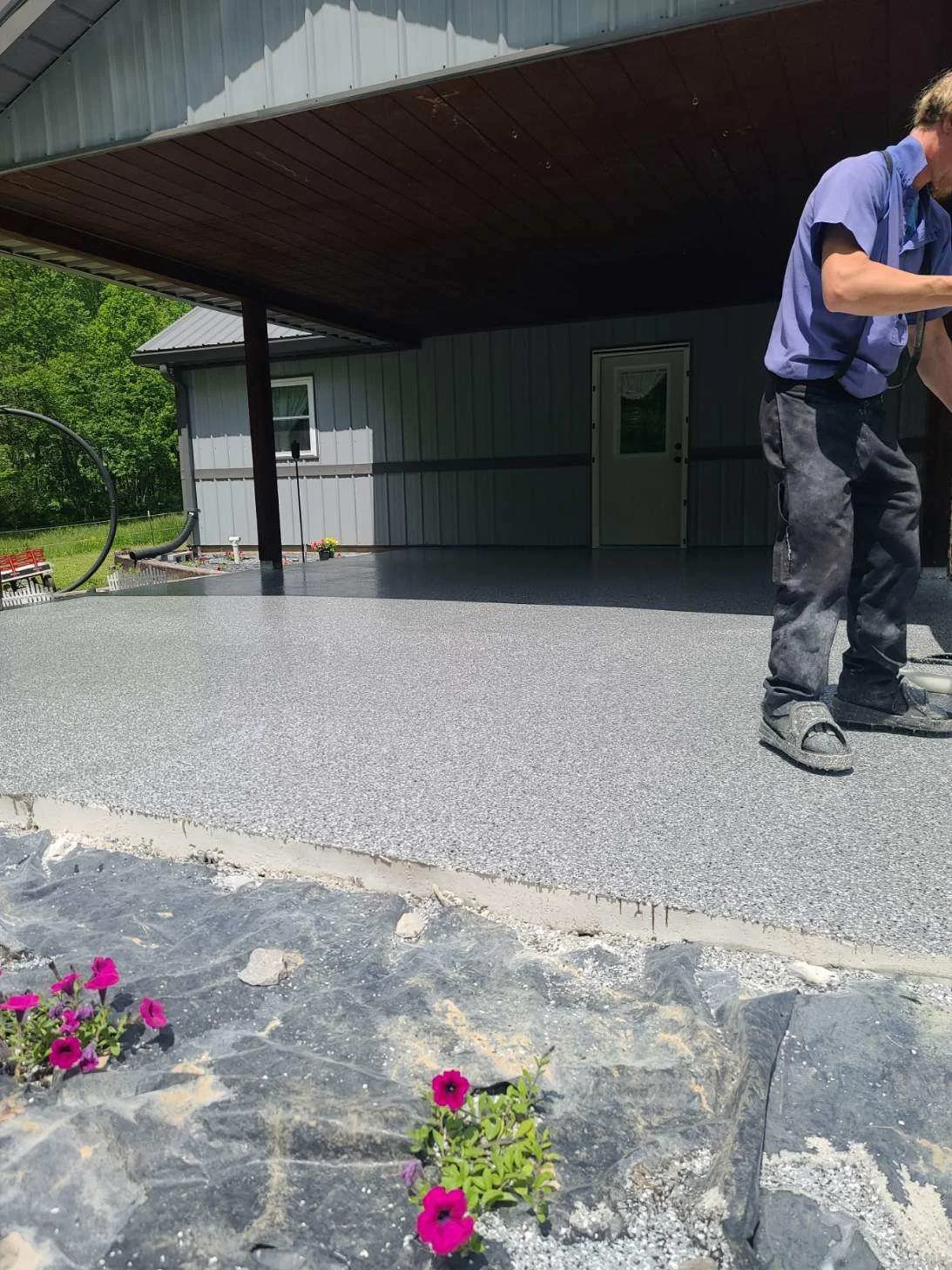 A person wearing a purple shirt and dark jeans is standing on a newly poured concrete patio with a textured surface, next to a house with a metal roof and siding. There are pink flowers in the foreground and green trees in the background.