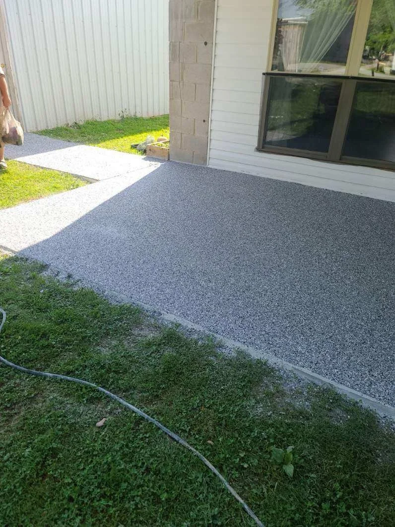 A newly poured concrete patio next to a house with white siding and a large window. A strip of grass surrounds the patio, with a garden hose in the lawn. Part of a person is visible on the left side.