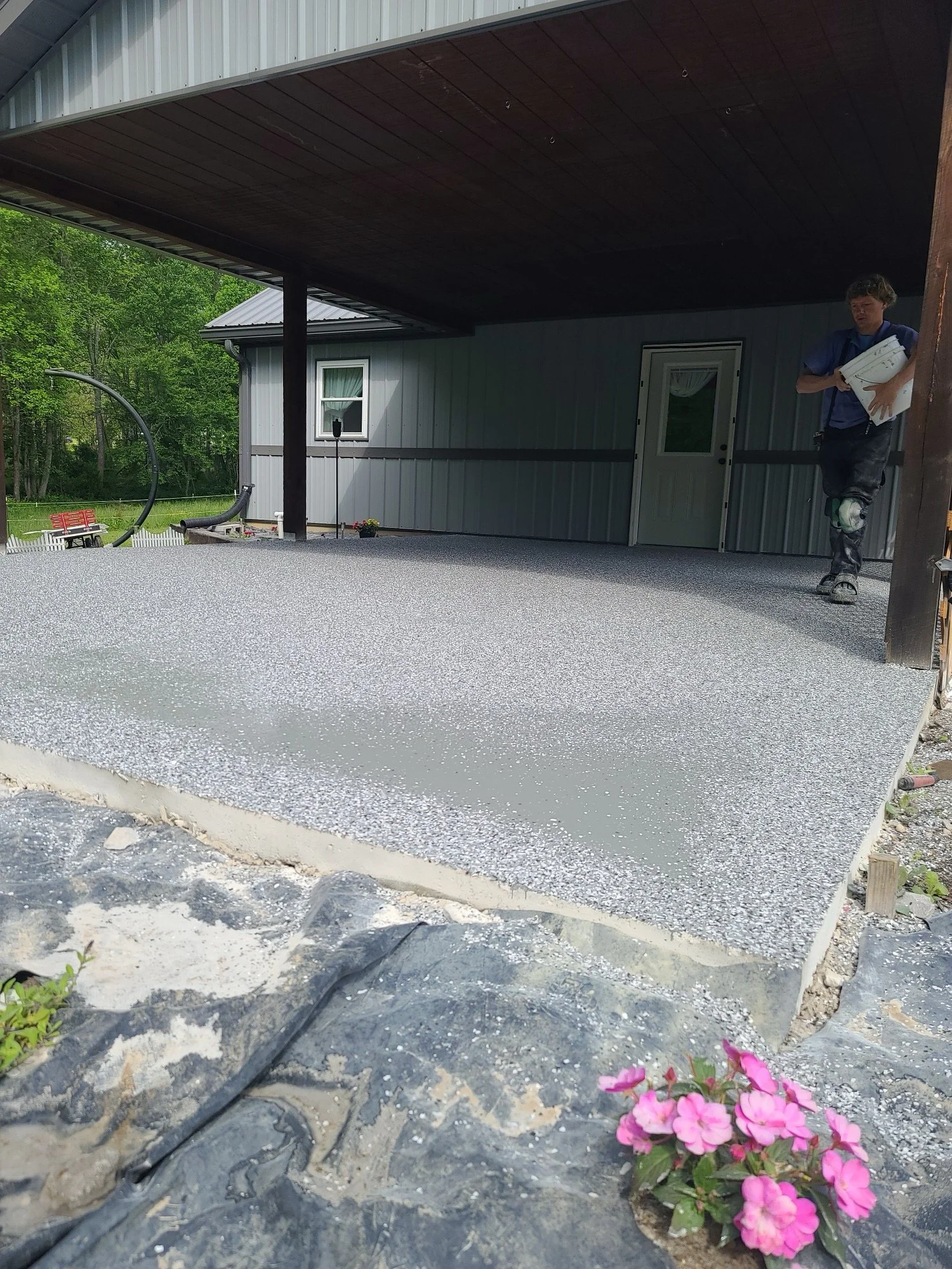 A construction worker with a knee brace holding a container of concrete on an elevated concrete porch behind a metal building. The porch has recently been poured and is still wet, with some gravel and a flower in the foreground.