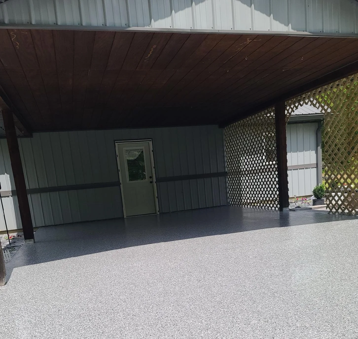 Covered porch area with a speckled gray concrete floor and gray metal siding, partially enclosed with lattice panels and a door leading inside.