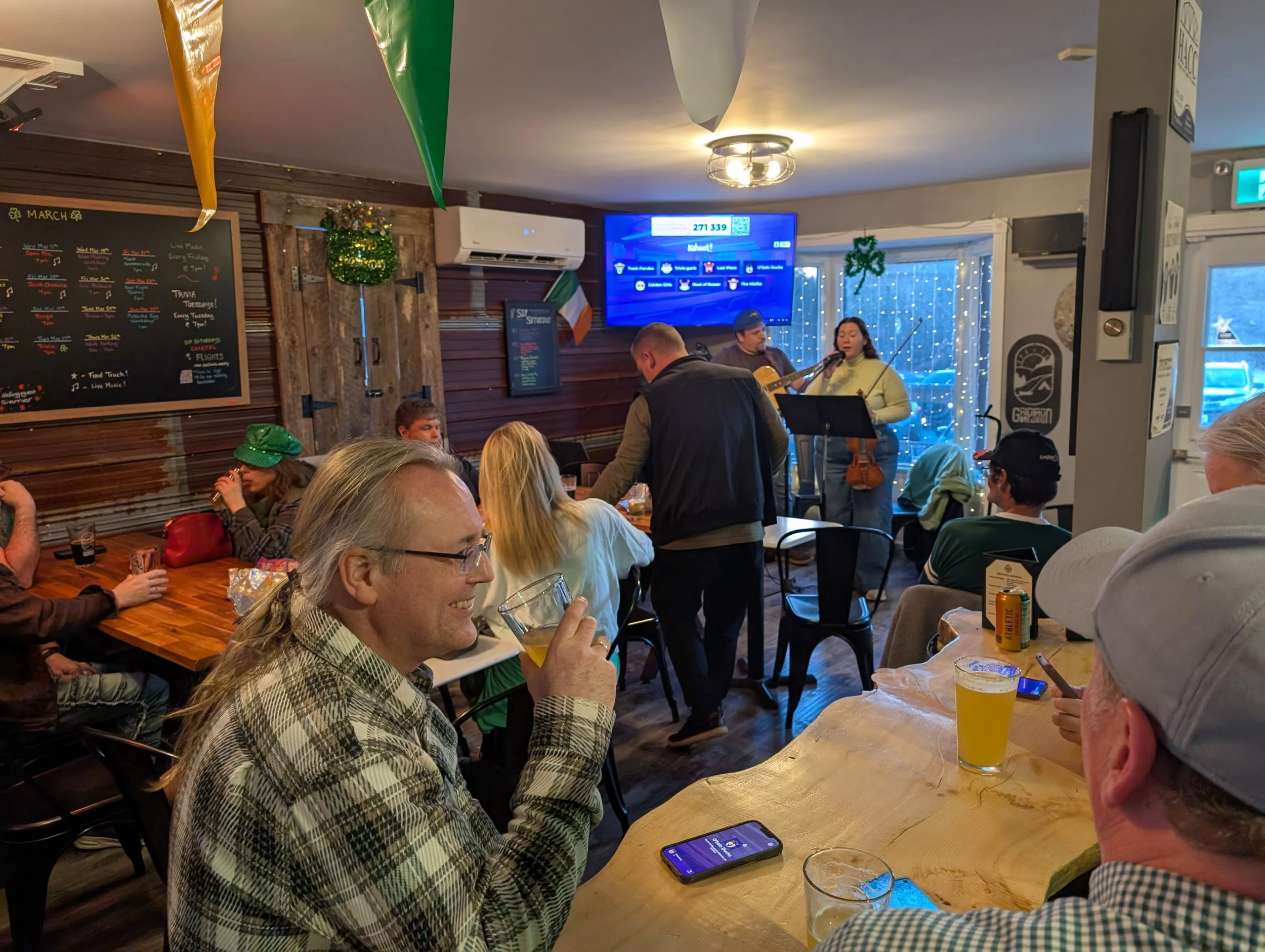 People enjoying live music performance in a cozy, decorated bar or pub, with some seated at tables and others at the bar, while a singer and guitarist perform on stage. The scene includes Christmas decorations and a visible menu on the wall.