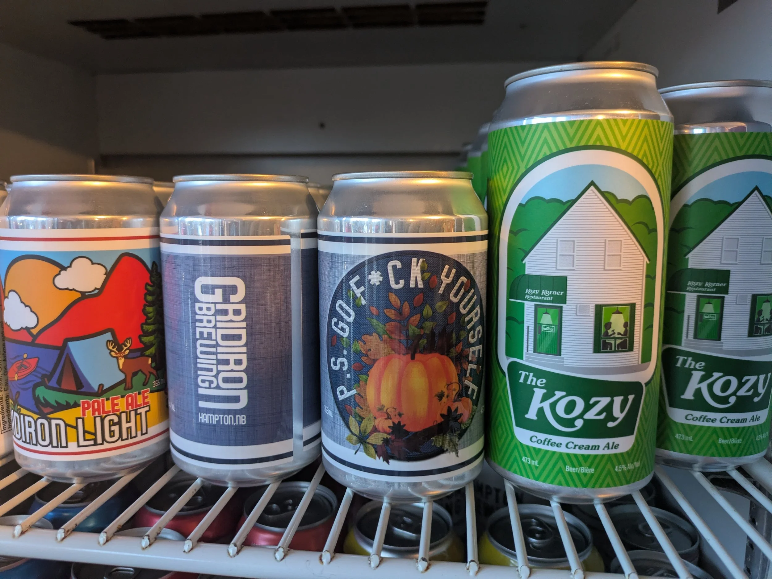 Cans of beer and a coffee-flavored ale on a refrigerator shelf. The beers include a Pale Ale Light, a Girdor G Brewing, a pumpkin-themed beer, and two cans of The Kozy Coffee Cream Ale.
