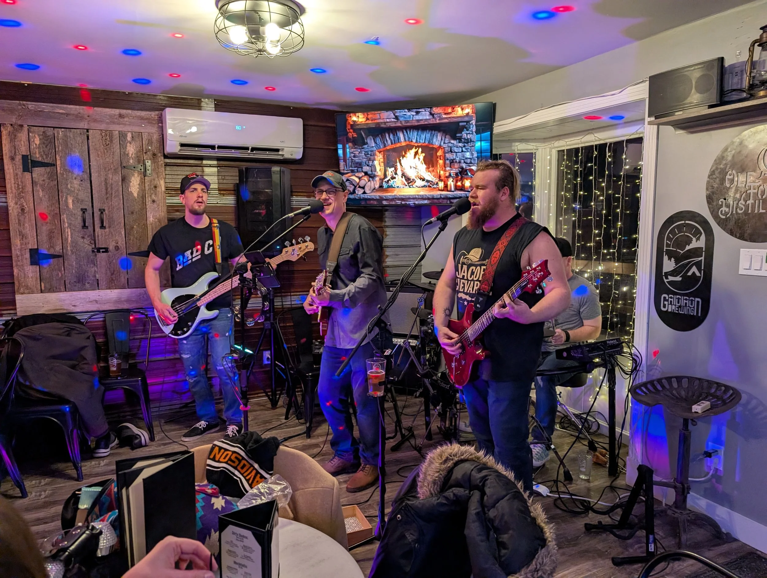 Three musicians playing guitars and singing at a bar, with a man on drums in the background. The scene is decorated with string lights and a framed picture of a fireplace on the wall.