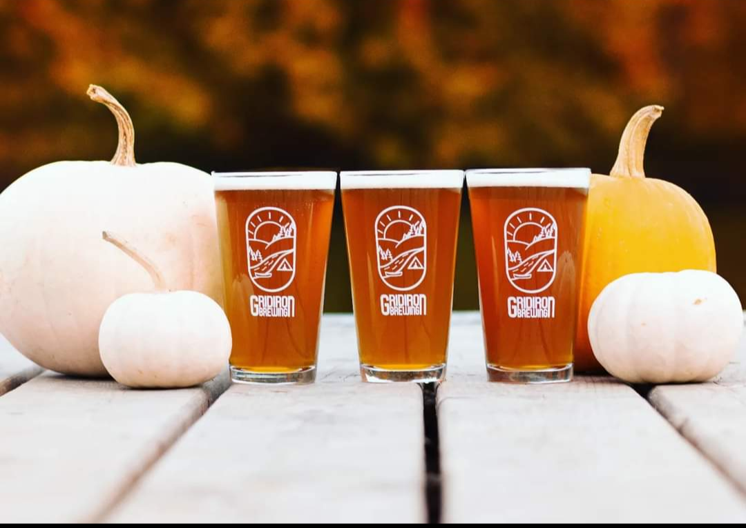 Three glasses of amber beer with a logo, flanked by white and orange pumpkins, on a wooden table outdoors with blurred fall foliage background.