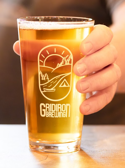 A person holding a glass of beer with a nature-themed logo and the text "Gridiron Brewing."