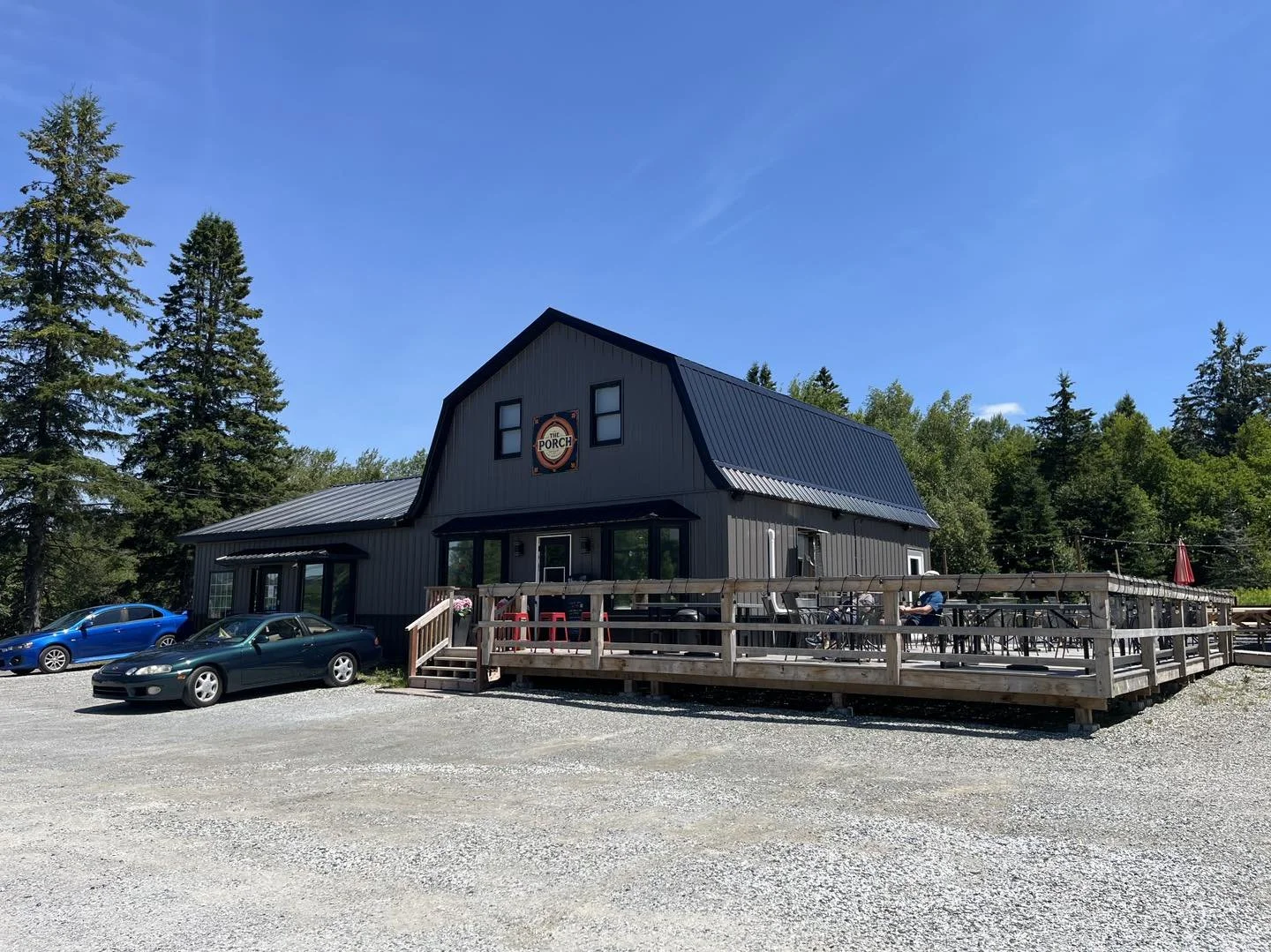 A black barn-style building with a sign reading 'The Porch' on the front, surrounded by trees and a gravel parking lot with parked cars, under a blue sky.