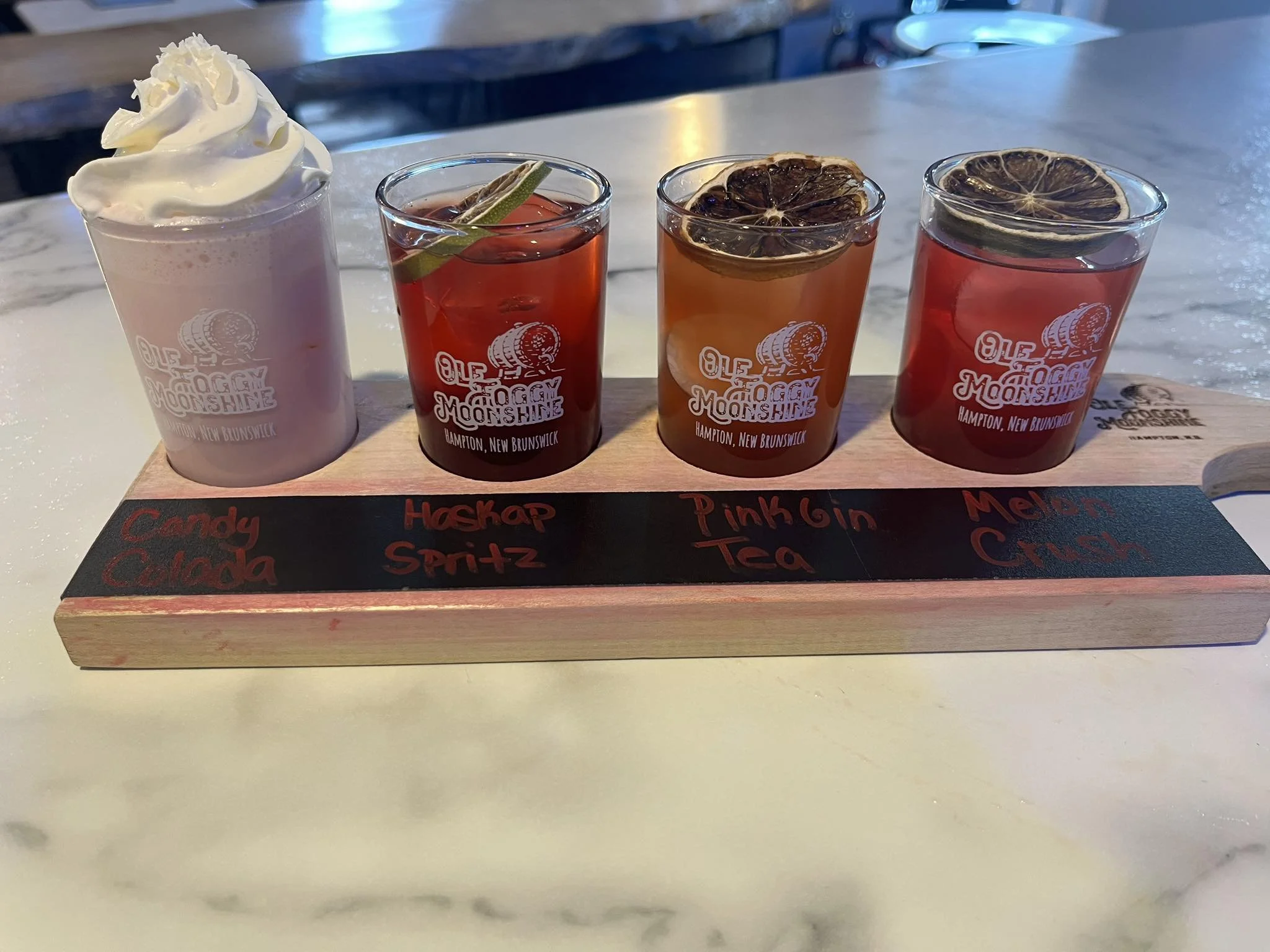 A wooden flight paddle with four small glasses of different colorful drinks, each garnished differently. The drinks are labeled Candy Calada, Haskap Spritz, Pink Gin Tea, and Melon Crush from left to right.