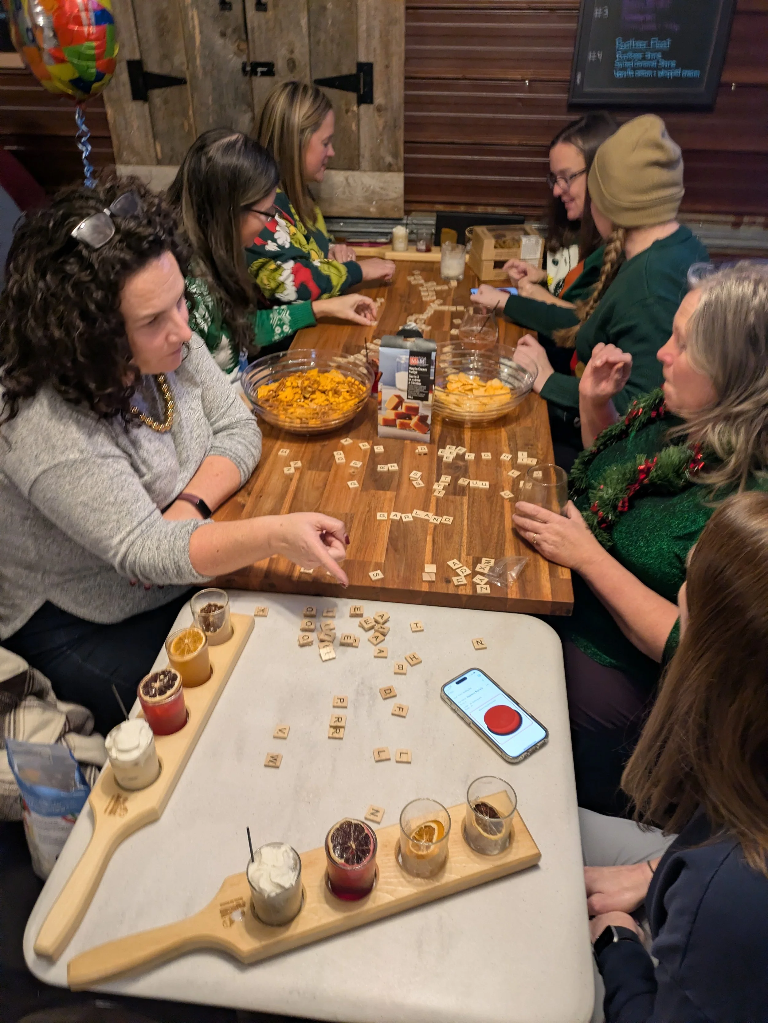 Group of women sitting around a wooden table playing a word game with Scrabble tiles and drinks, in a rustic indoor setting with wood-paneled walls and a menu board.