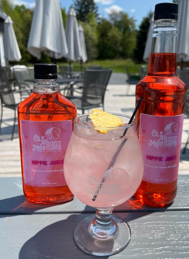 Two bottles of Hippie Juice with pink labels and a glass of pink drink with a pineapple wedge on top, set outdoors on a gray table with umbrellas and chairs in the background.