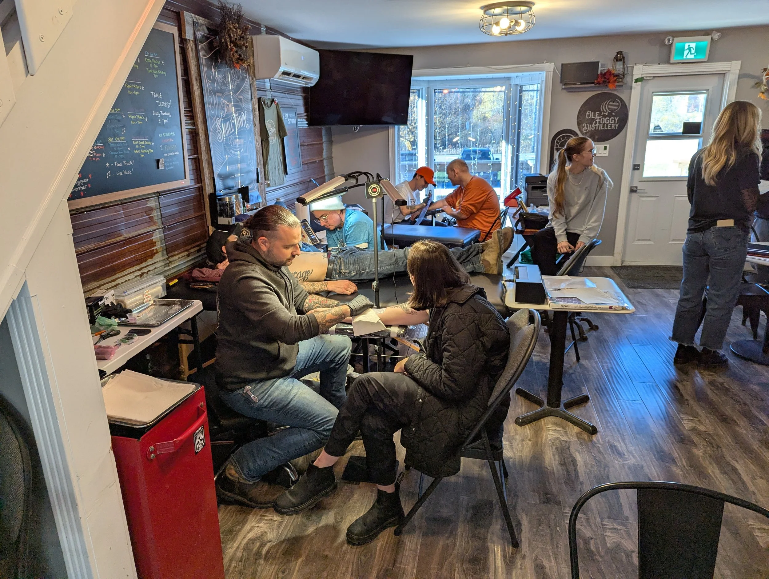Tattoo artist in black hoodie tattooing a woman's forearm in a cozy tattoo shop with dark wood floors, wall decor, and several clients getting tattoos.