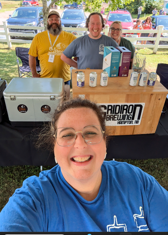 A woman with glasses and a blue shirt taking a selfie with three smiling people behind her at a booth for Cardiron Brewing in Hampton, NB. The booth has canned drinks, cartons, and a cooler, with a wooden sign displaying the brewery's name.