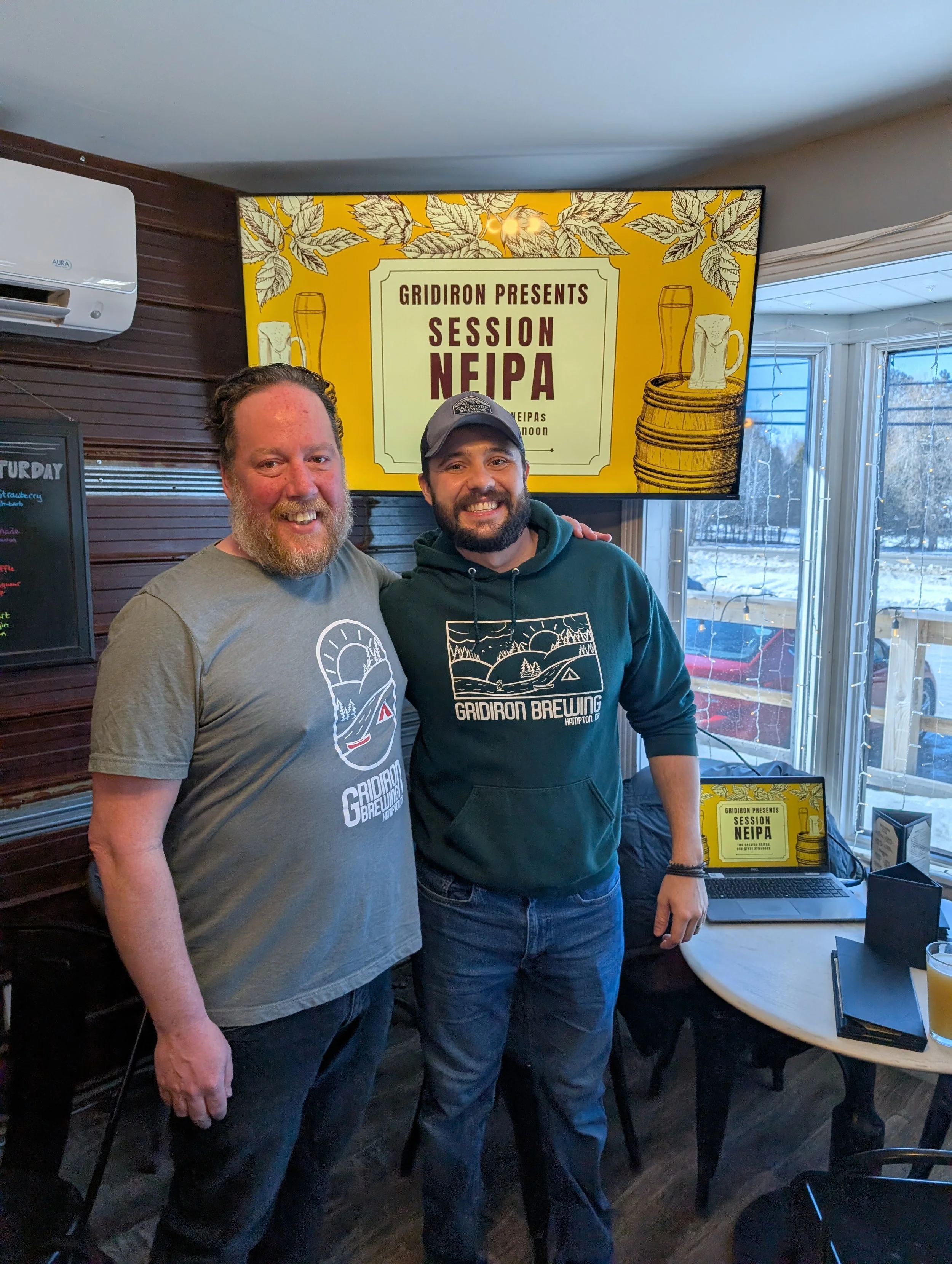 Two men standing indoors, smiling, with a large screen behind them displaying a sign that reads 'Gridiron Presents Session NEIPA.' One is wearing a gray T-shirt with a logo, and the other is wearing a green hoodie with a logo. There is a window with 