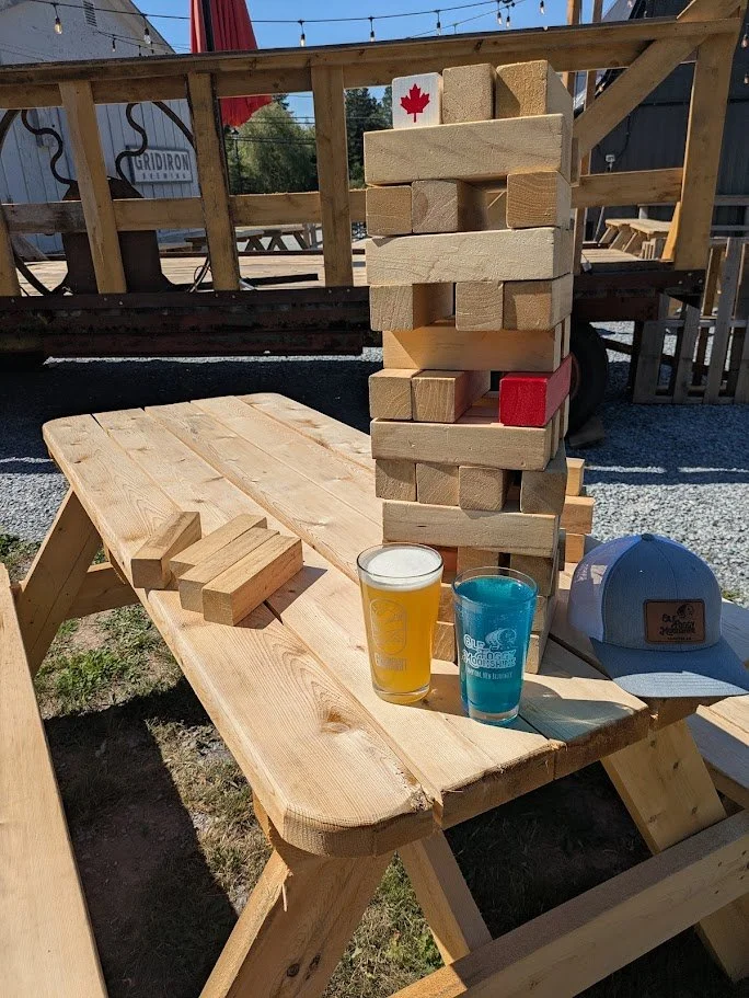 A large wooden Jenga game on a picnic table with a glass of beer, a blue cocktail, a gray baseball cap, and a small wooden block beside it. In the background, there is a wooden deck and string lights, indicating an outdoor setting.