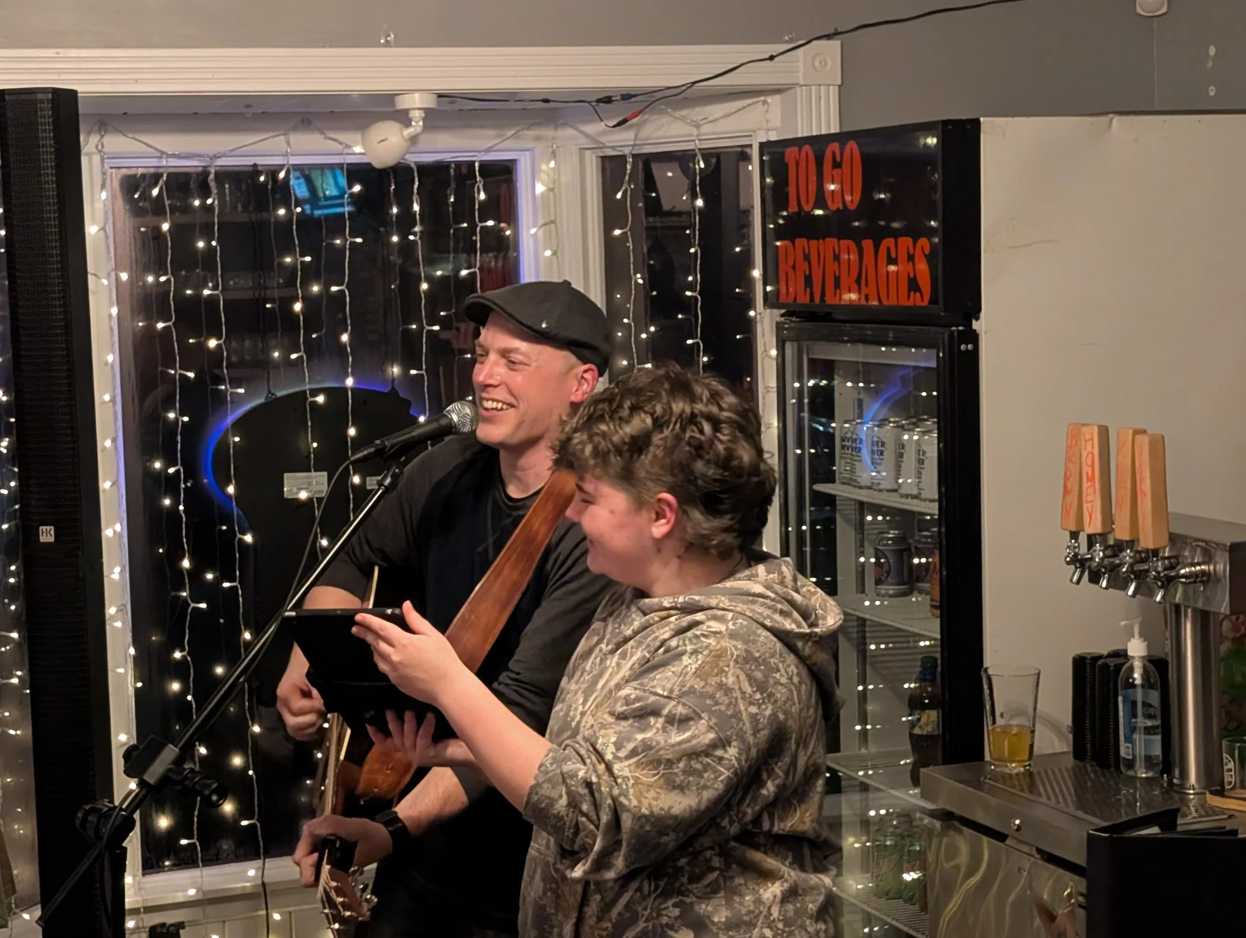 Two men singing and playing guitars at a bar or pub with festive string lights in the background and a sign that says 'To Go Beverages'.