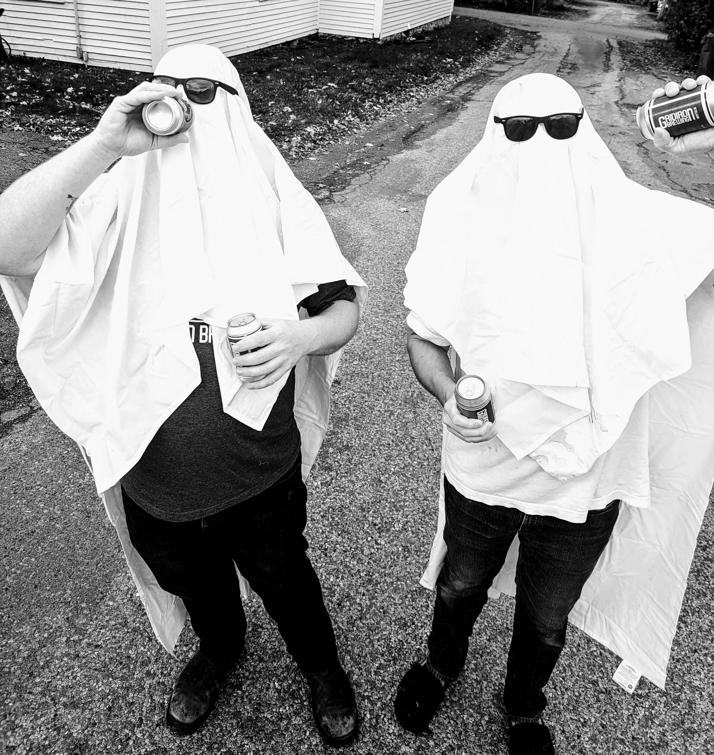 Two people standing outside, wearing white cloths over their heads and sunglasses, holding cans of drinks, with one person drinking from a can and the other holding a can in both hands.