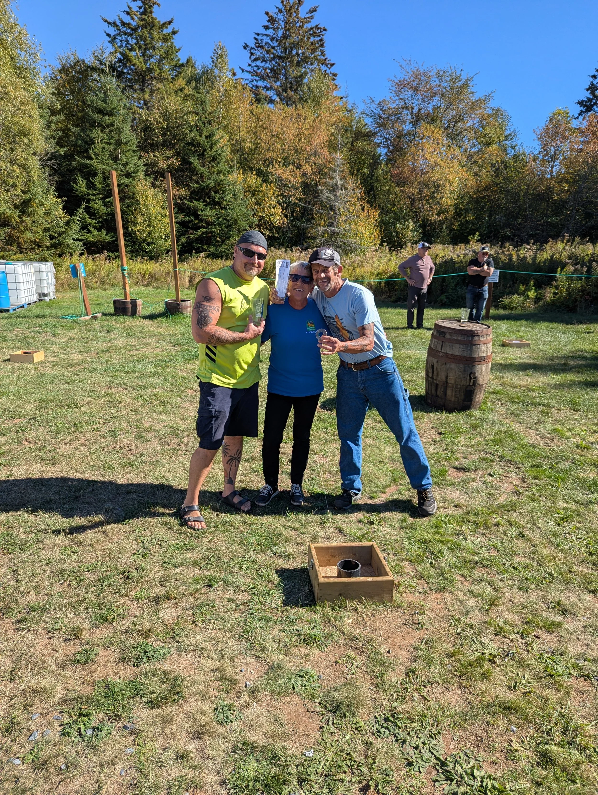 A group of three people standing outdoors on a sunny day, smiling and holding awards or prizes, with a grassy field, trees, and other people in the background. Playing washer toss. 