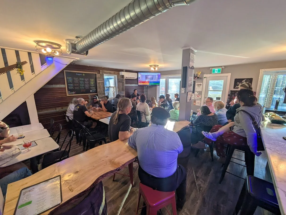 Inside a busy restaurant with people dining at tables and a bar area, TV screens on the wall, and a chalkboard menu.