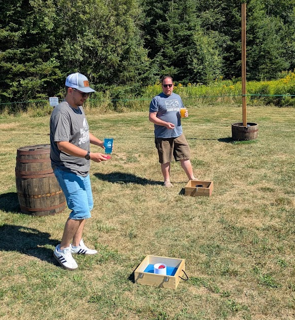 Two men playing cornhole outdoors on a grassy field with trees in the background. One man is in the foreground, wearing a gray shirt, shorts, and a baseball cap, holding a blue cup. The other man is in the background, wearing sunglasses, a blue t-shi