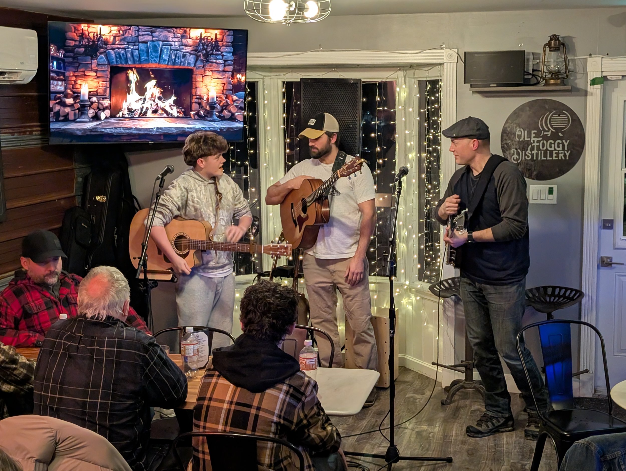 Three musicians performing with acoustic guitars on a small stage in a cozy, decorated venue, audience seated at tables in the foreground, with fairy lights and a large fireplace graphic on a TV screen in the background.