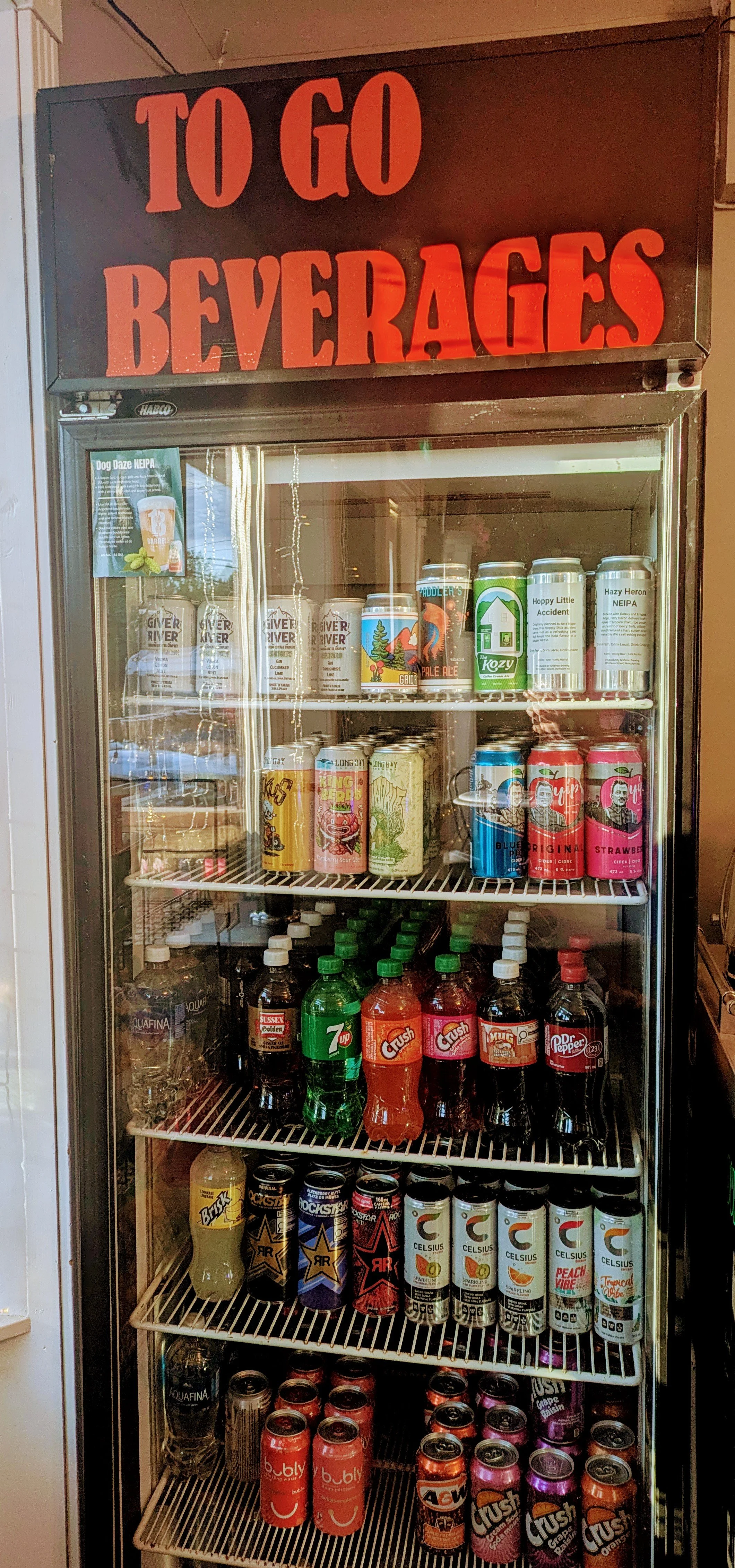 A refrigerator filled with various canned and bottled beverages including soda, energy drinks, and craft beers, with a sign on top reading 'TO GO BEVERAGES' in large red letters.