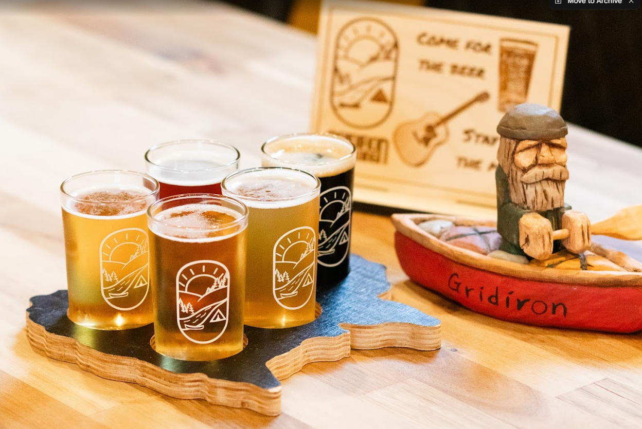 Four glasses of beer on a wooden table, with a decorative board and a wooden boat with a carved figure, labeled 'Gridiron'.