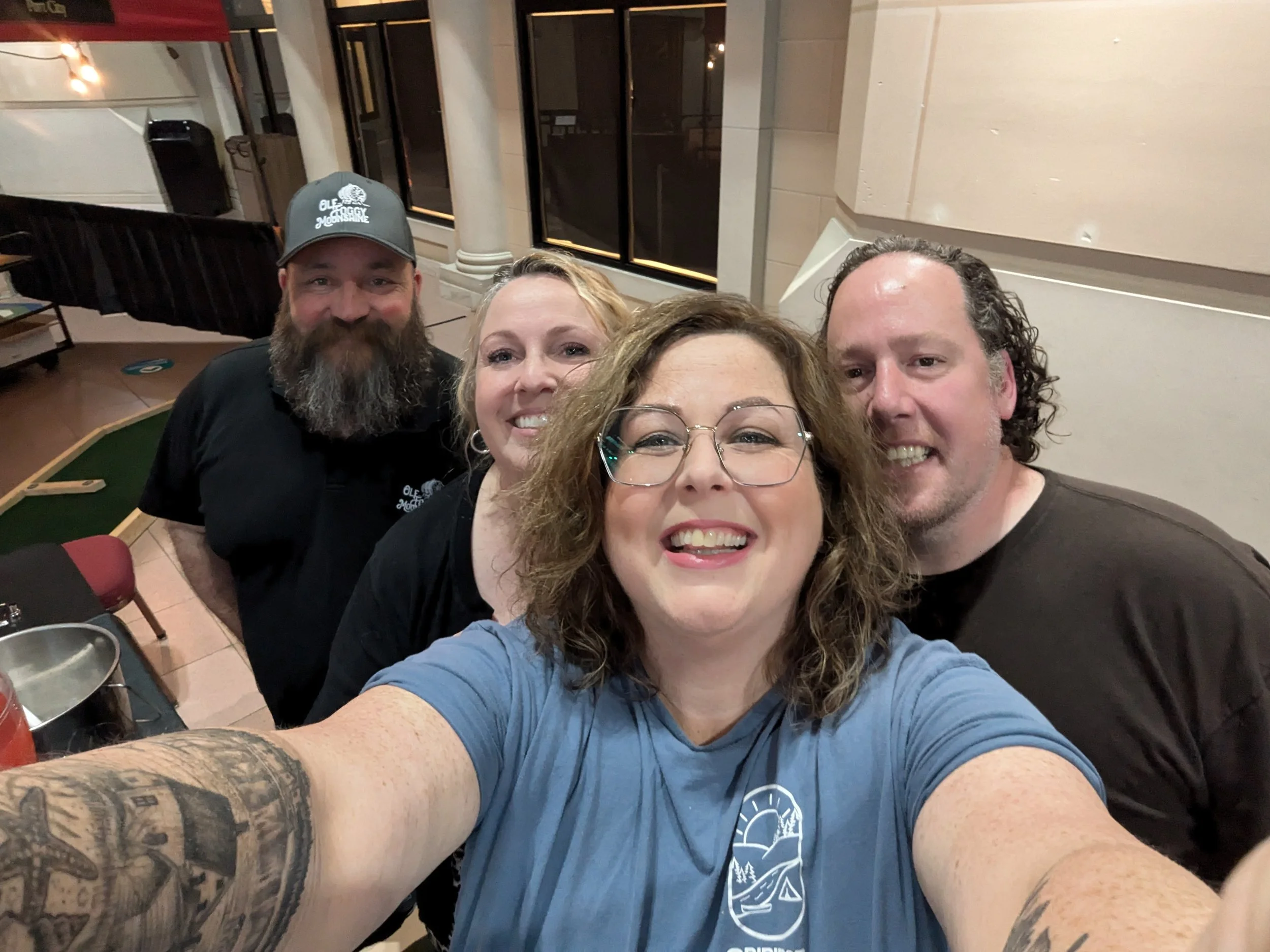 Group of four smiling people taking a selfie indoors. Woman in front with glasses, wearing a blue shirt with a logo. Man on right with curly hair, wearing a dark shirt. Woman behind with blonde hair, smiling. Man on left with beard, wearing a black s