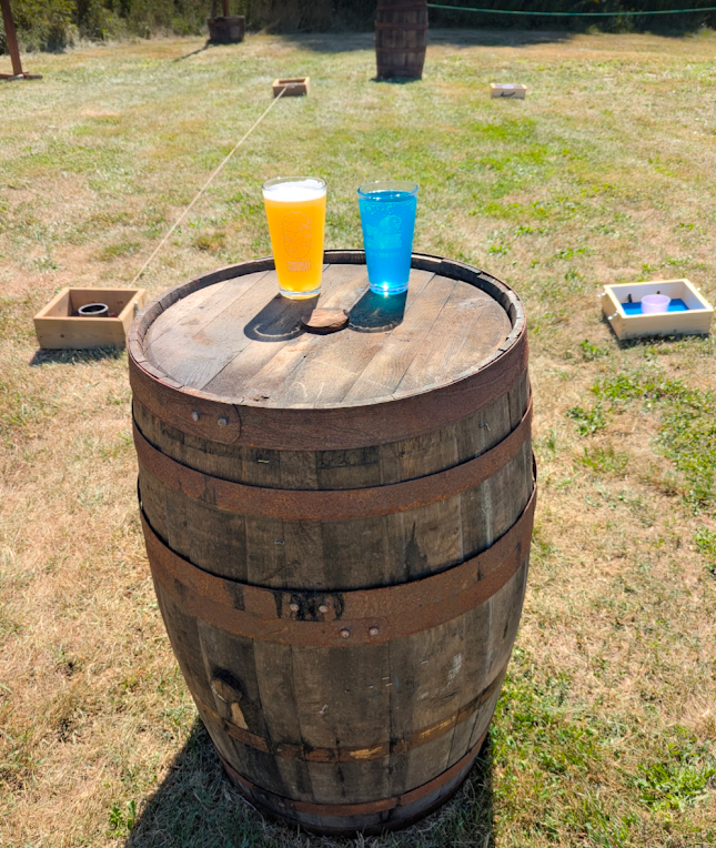 Two glasses, one orange and one blue, are placed on top of a wooden barrel outdoors. The orange glass is filled with a yellow beverage, and the blue glass is empty. In the background, there are two cornhole game boards on a grassy lawn and a tree tru