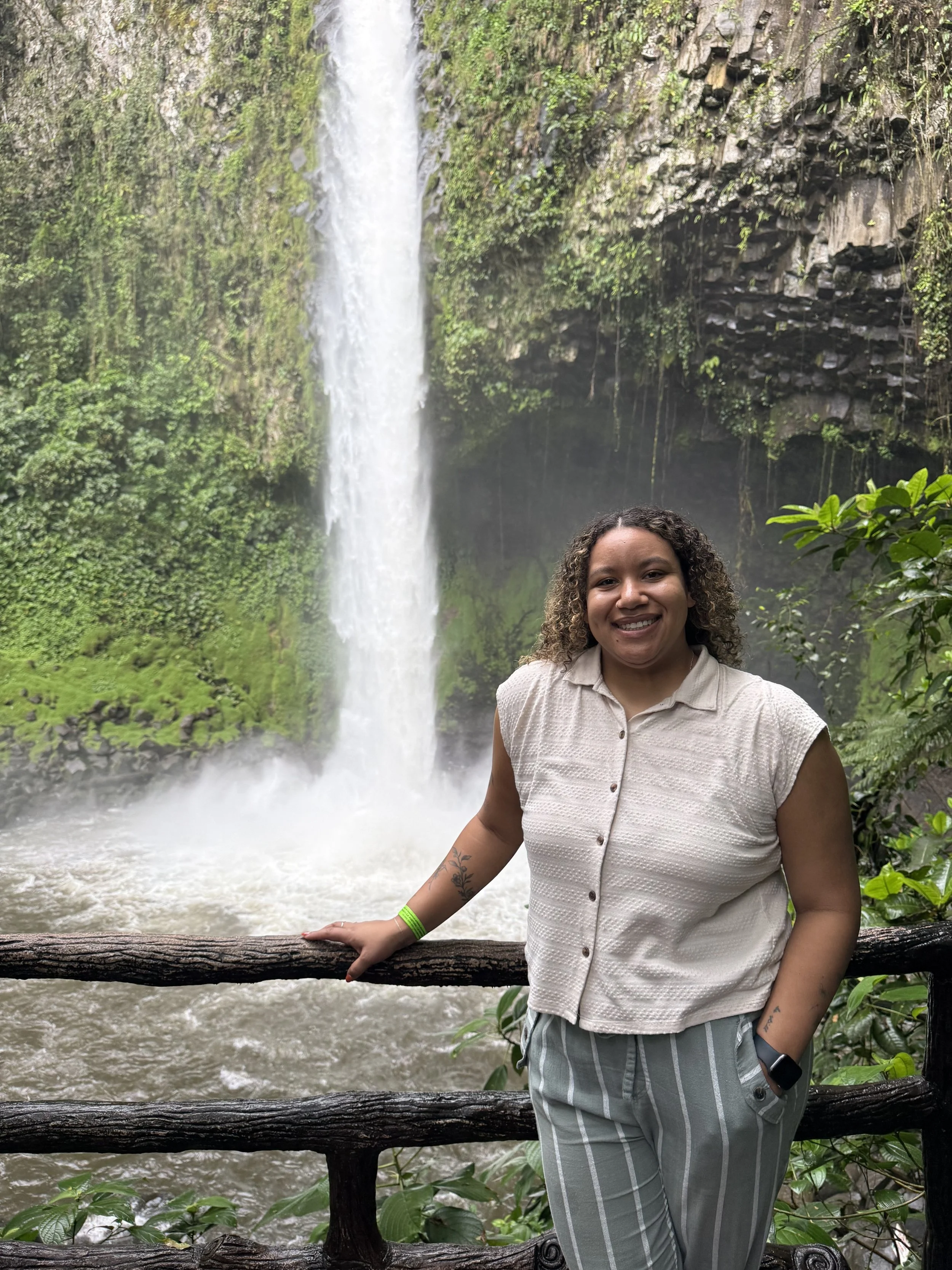 A woman with curly hair smiling at the camera in front of a waterfall surrounded by lush greenery.