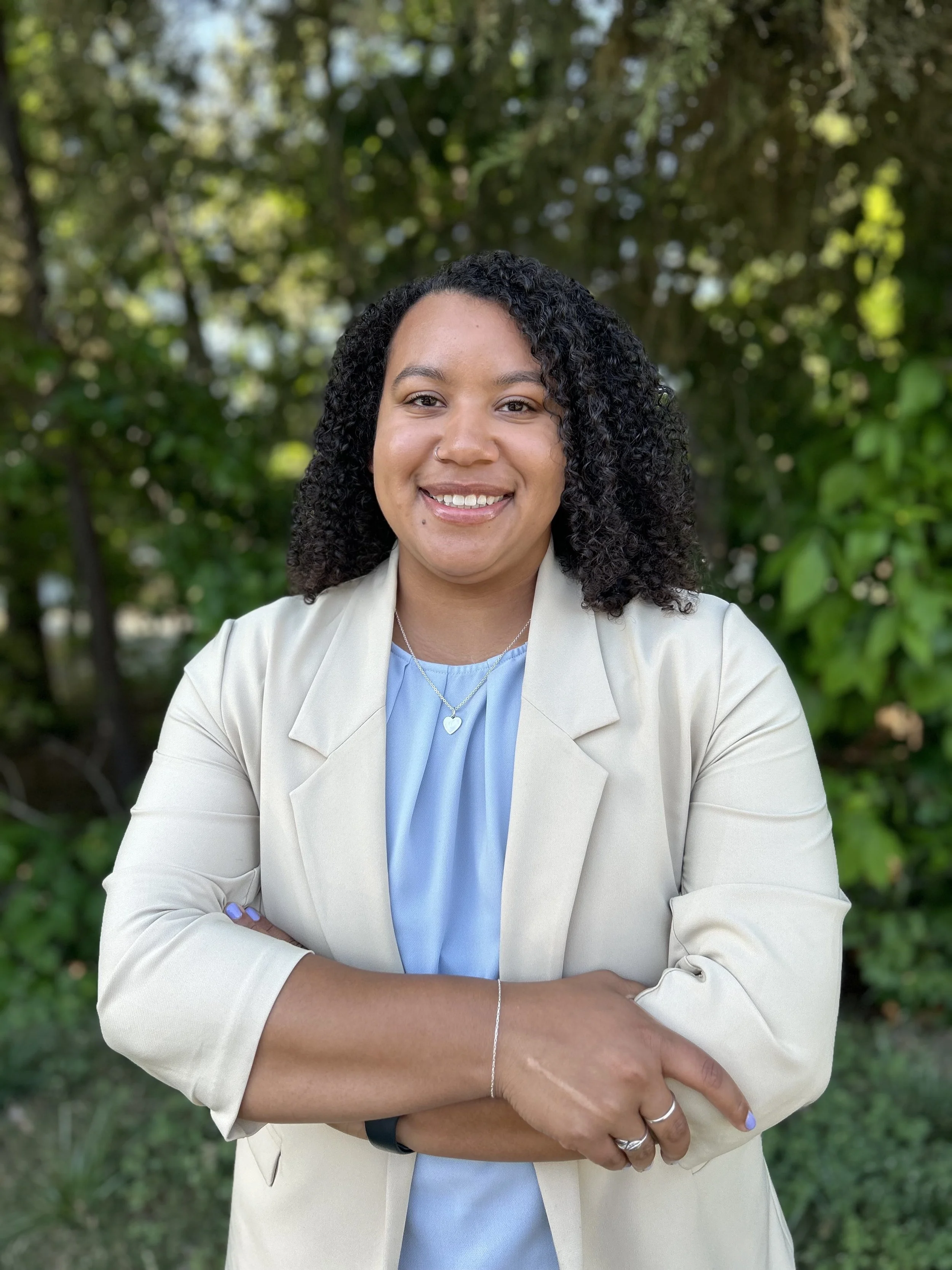A woman with curly black hair, wearing a beige blazer over a light blue top, smiling with arms crossed, standing outdoors with green trees and foliage in the background.