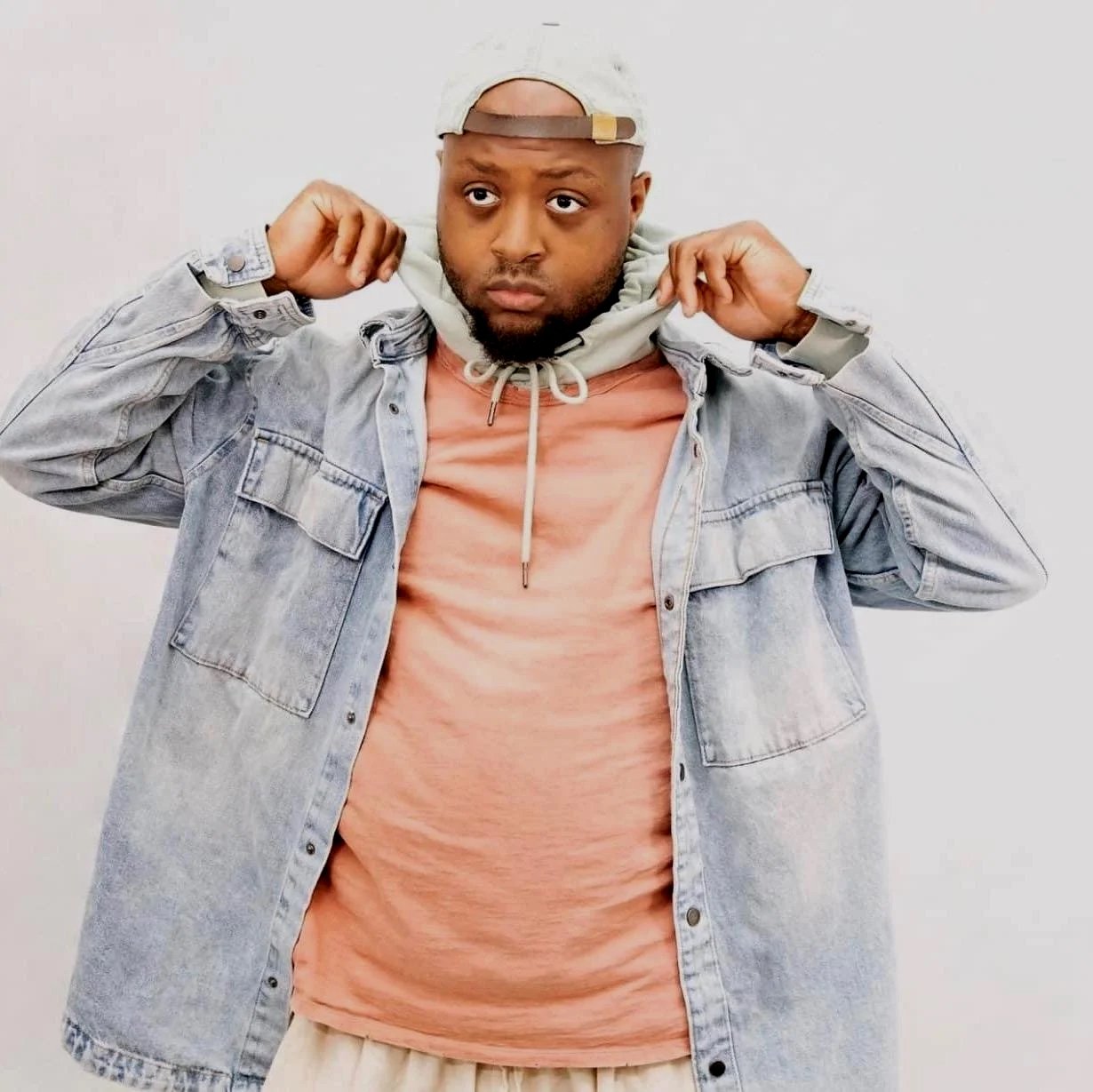 A man in a light denim jacket, peach t-shirt, and beige pants, wearing a backward cap and hoodie, adjusting the hoodie around his neck.