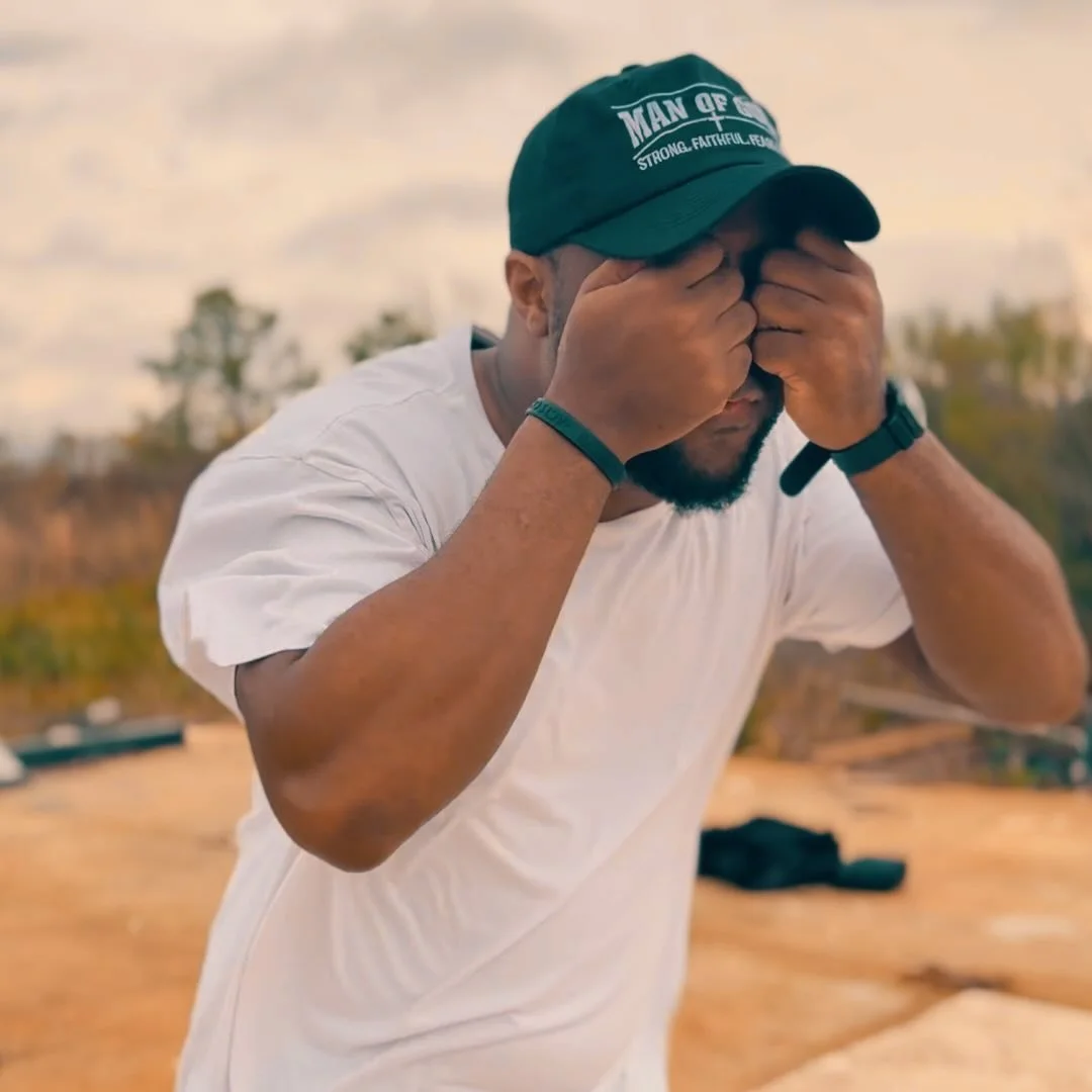 A man wearing a green cap and white t-shirt covers his face with both hands outdoors during daytime.